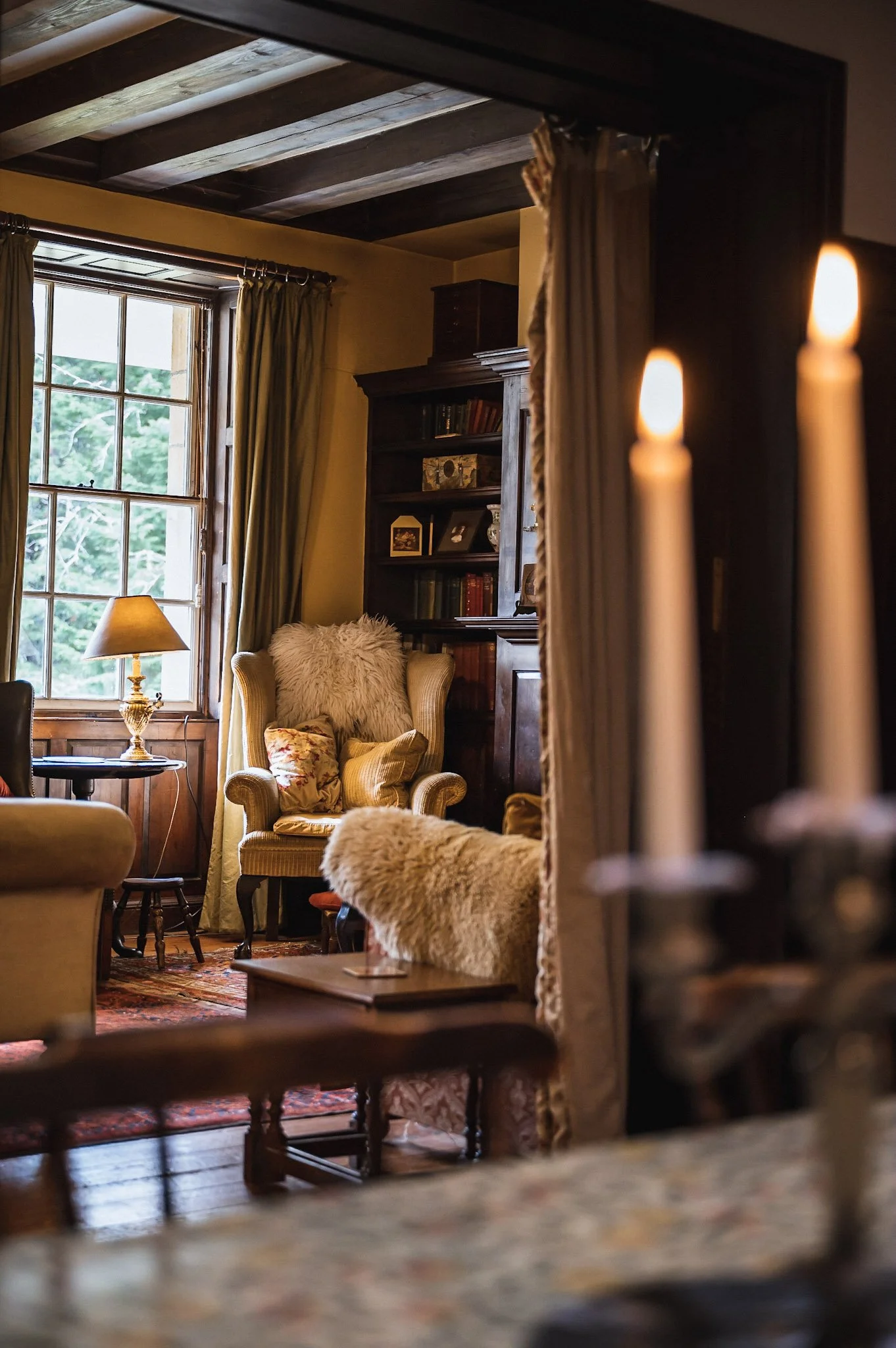 Cozy living room with a large window, yellow armchair with pillows, bookshelf, table lamp, and curtains, framed by a doorway with candles in the foreground.