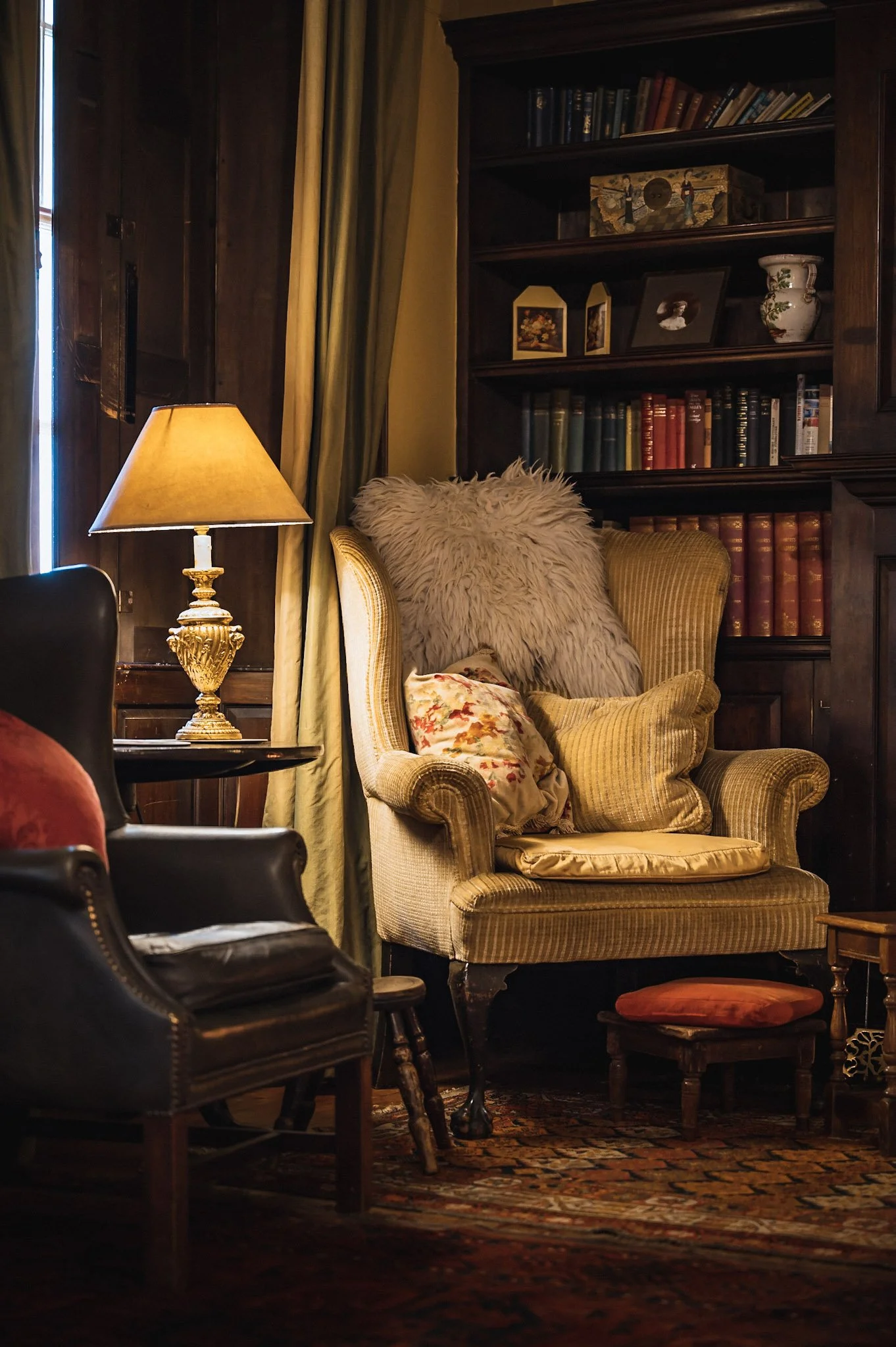 A cozy living room with a beige armchair decorated with fluffy pillows, a leather armchair, a side table with a gold ornate lamp, and a wooden bookshelf filled with books and decorative items.