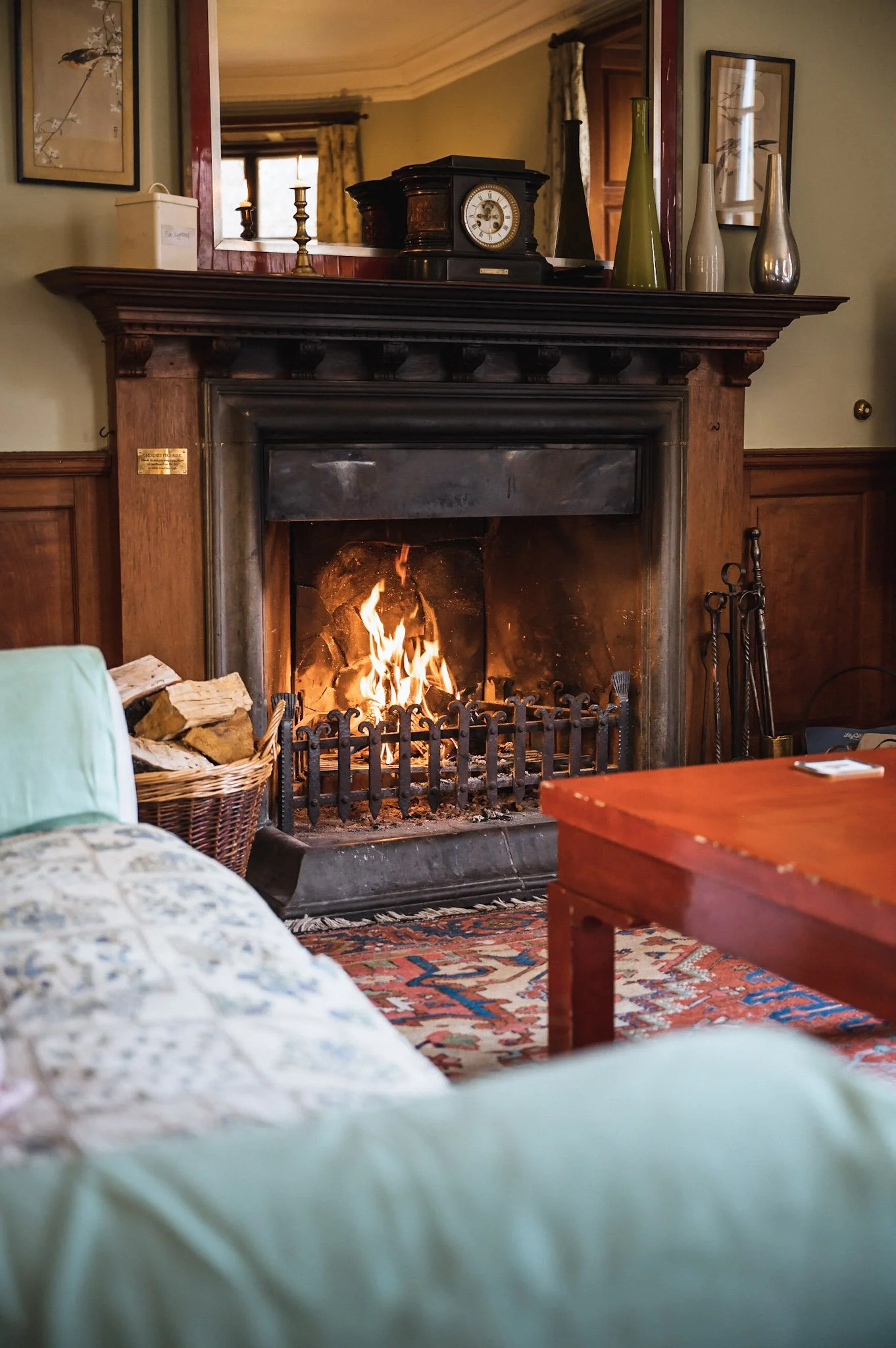 A cozy living room with a lit fireplace, a basket of firewood on the left, a wooden coffee table in the foreground, and a patterned rug. Decorative vases and a clock are on the mantel above the fireplace, with framed artwork and a mirror on the walls.