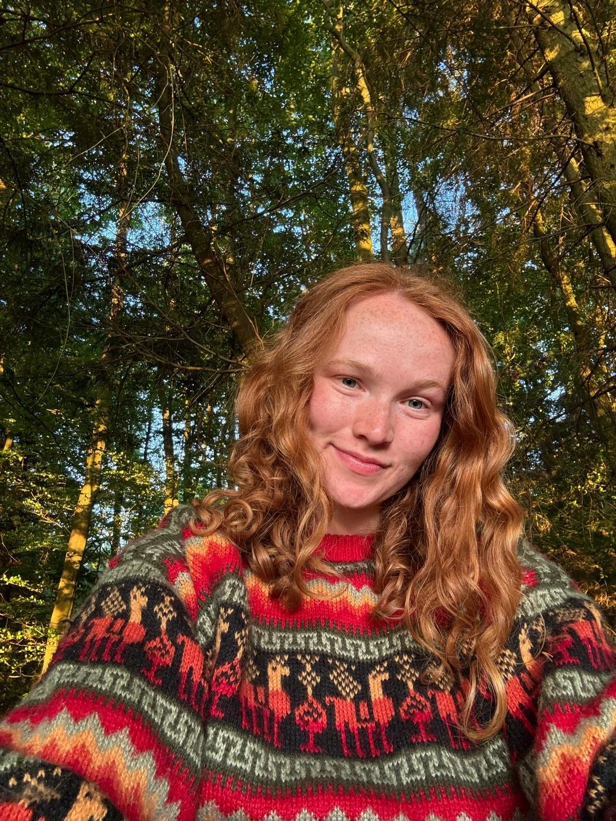 A young woman with curly red hair and freckles smiling outdoors among trees, wearing a colorful, patterned sweater.