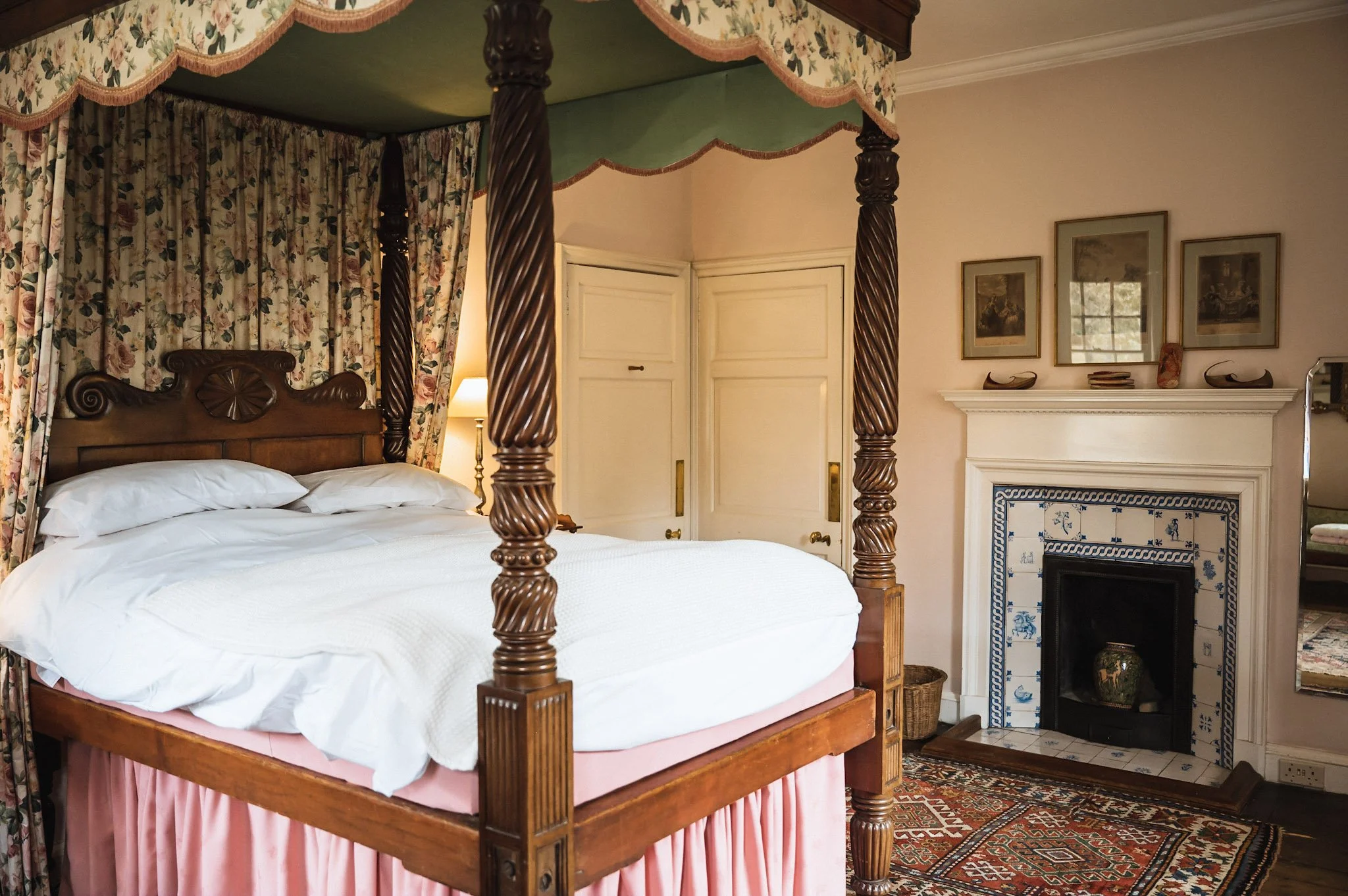 A vintage bedroom with a four-poster wooden bed with carved posts, white bedding, colorful drapes, a decorative fireplace with blue and white tiles, framed artwork on the wall, a mirror, and a Persian rug.
