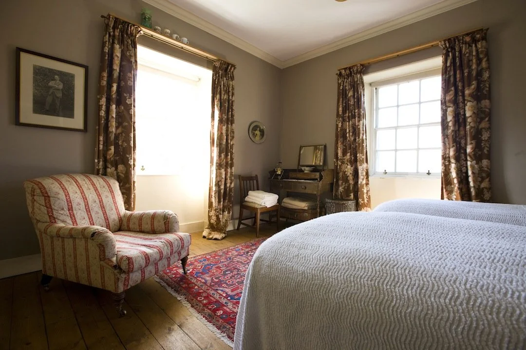 A cozy bedroom with two large windows draped with floral curtains, a vintage armchair with red and yellow patterns, a wooden desk with a mirror, and a bed with a white textured bedspread. An area rug lies on the wooden floor, and framed pictures hang on the walls.