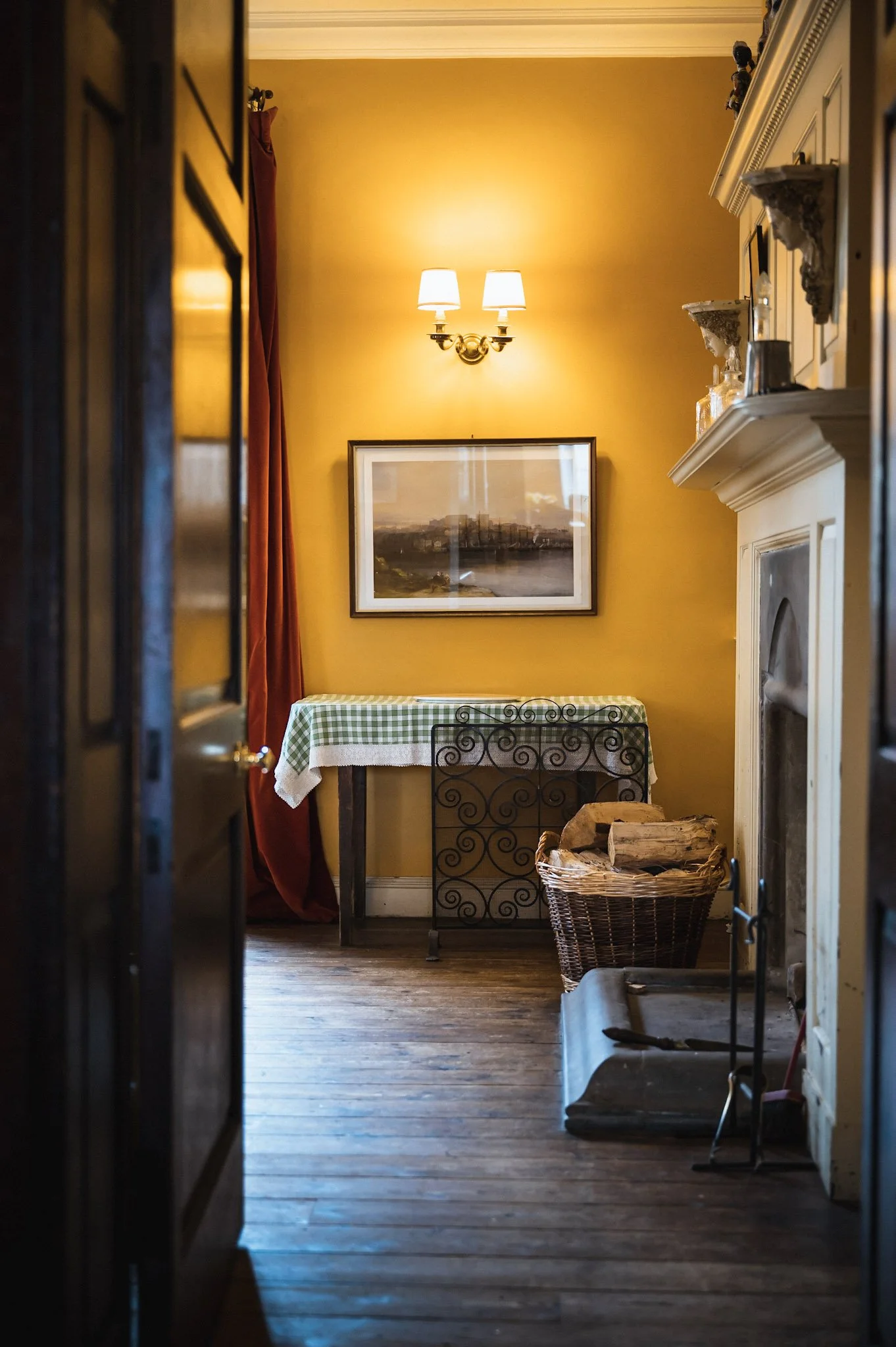 Interior of a cozy room featuring a yellow wall, a framed landscape painting, a table with a green checkered cloth, a wrought iron chair, a basket of firewood, a fireplace, and a vintage chandelier with four thumbs-up shades.
