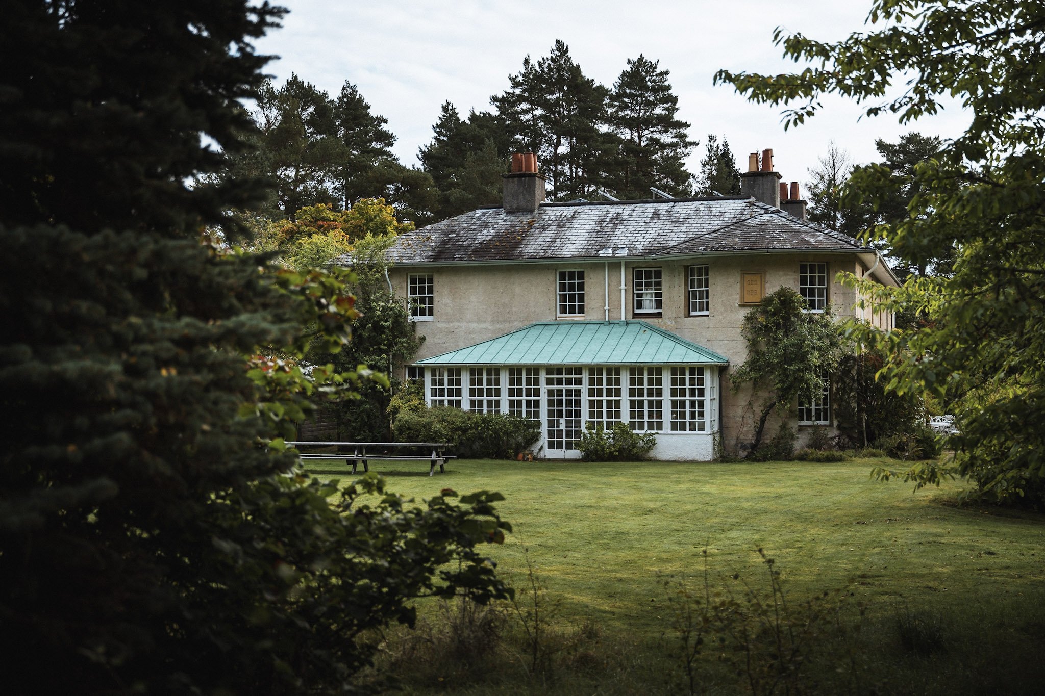A large house with a weathered roof, multiple chimneys, and a prominent glass-enclosed sunroom, surrounded by lush greenery and trees.