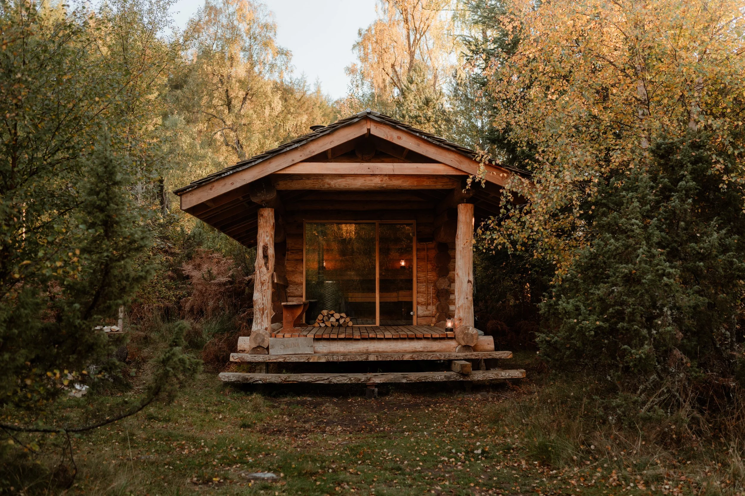 A small wooden cabin with a porch, surrounded by autumn trees, with logs stacked on the porch and the interior softly lit visible through a glass door.