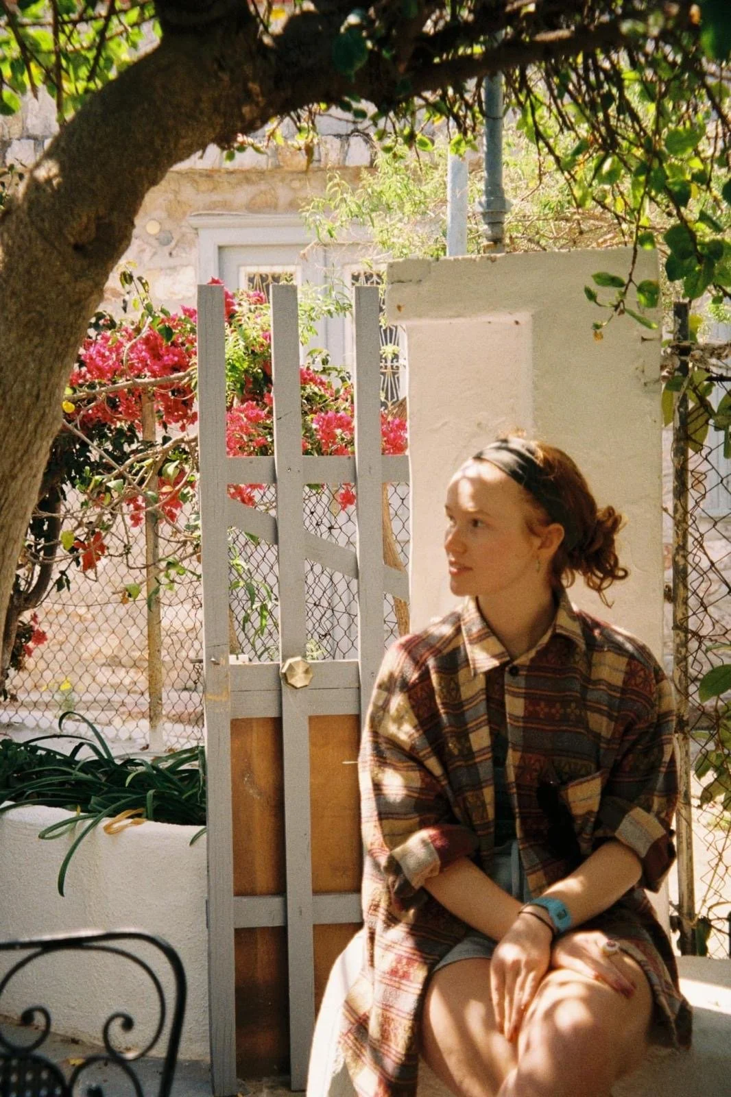 A young woman sitting outdoors under a tree, wearing a plaid shirt and shorts, with sunlight and shadows on her face and legs, and pink flowers in the background.