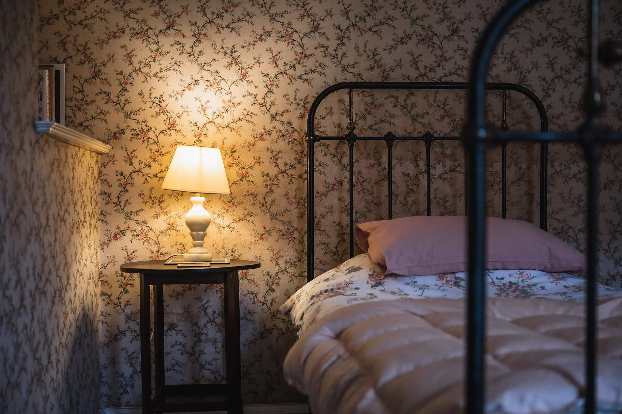 A cozy bedroom with floral wallpaper, a black metal bed frame, pink pillow, and a bedside table with a lit lamp.