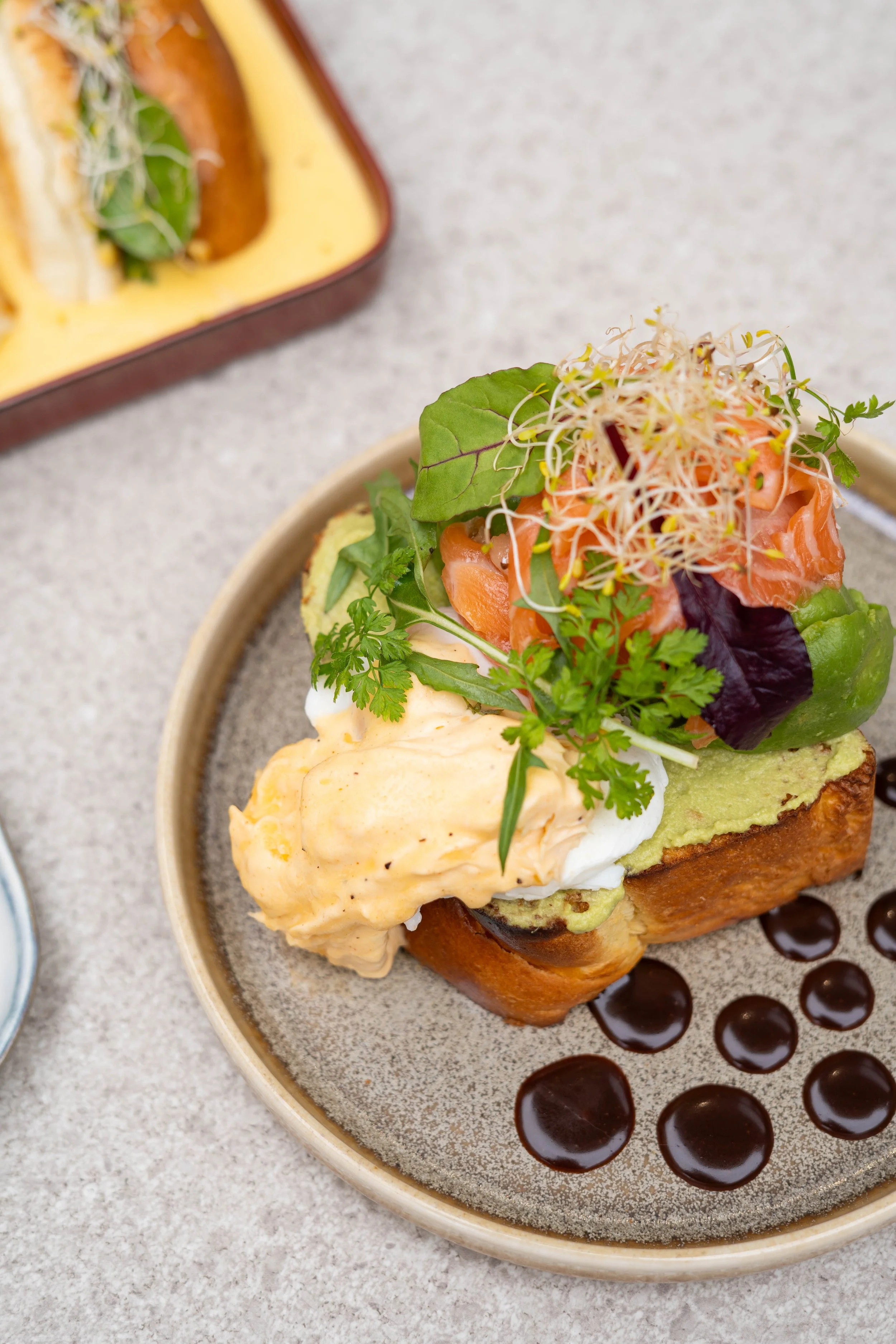 Oeufs bénédictine avec saumon fumé, avocat, jeunes pousses et sauce hollandaise sur pain grilled, décoré de coriandre, avec un dessin de sauce chocolat en cercle dessous
