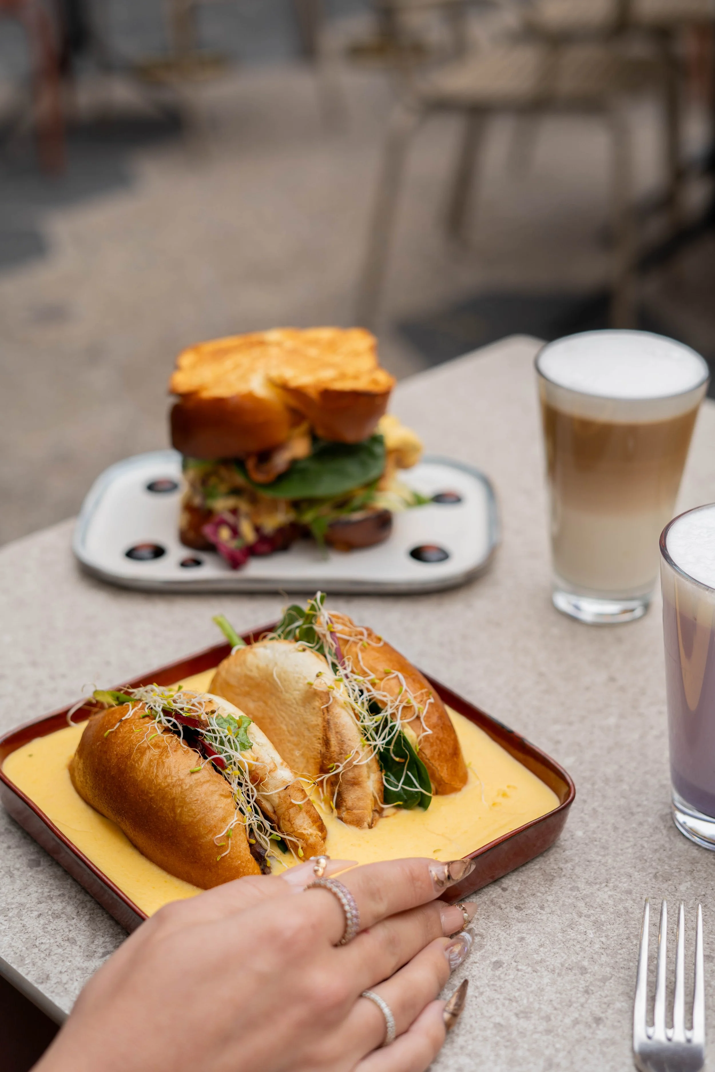Assiette de sandwichs avec une sauce crémeuse et garnitures végétales, accompagnée de deux boissons à lattes avec mousse et d'un sandwich en arrière-plan sur une table extérieure.