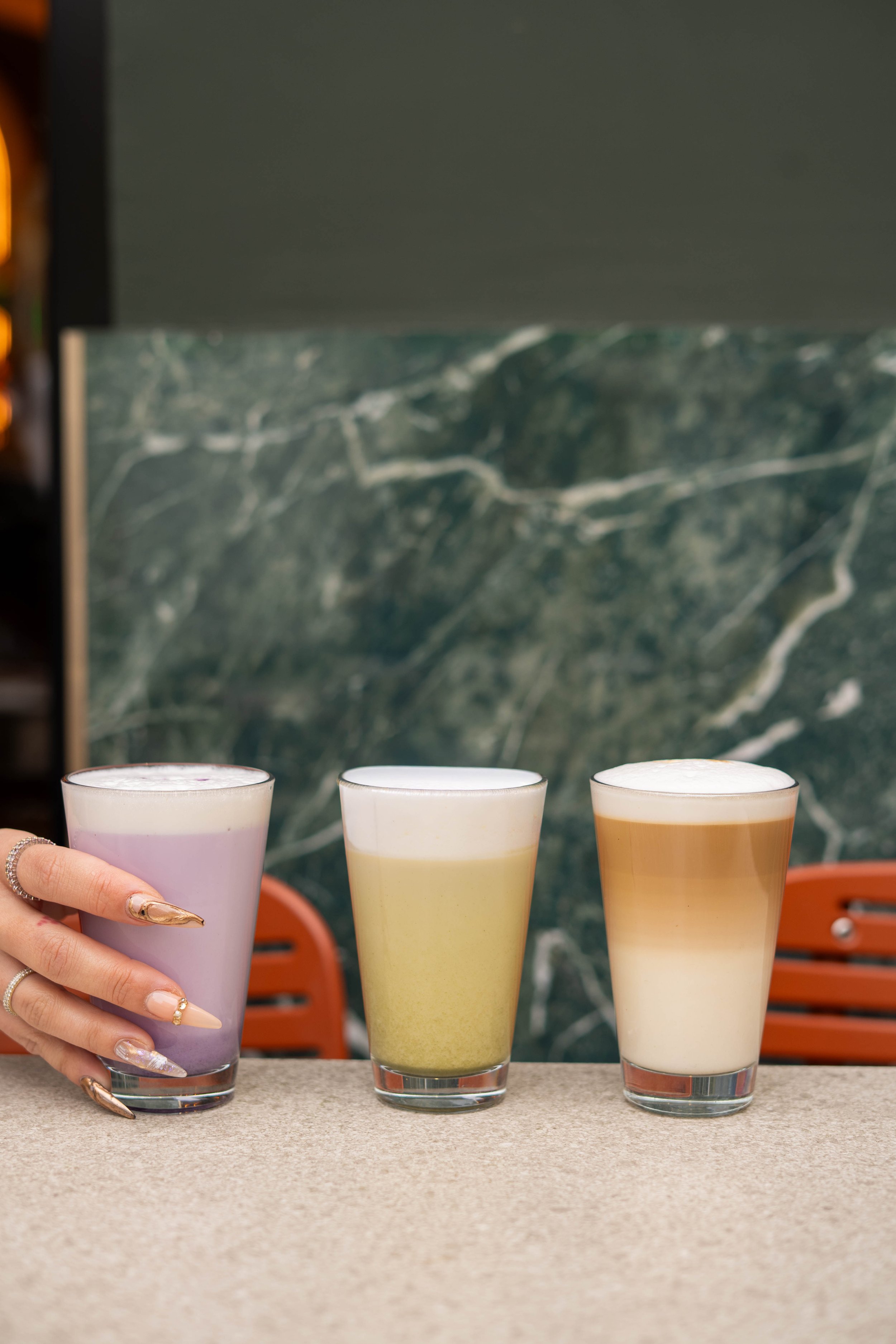 Trois verres de boissons colorées sur une table, avec une main portant un verre violet à gauche.