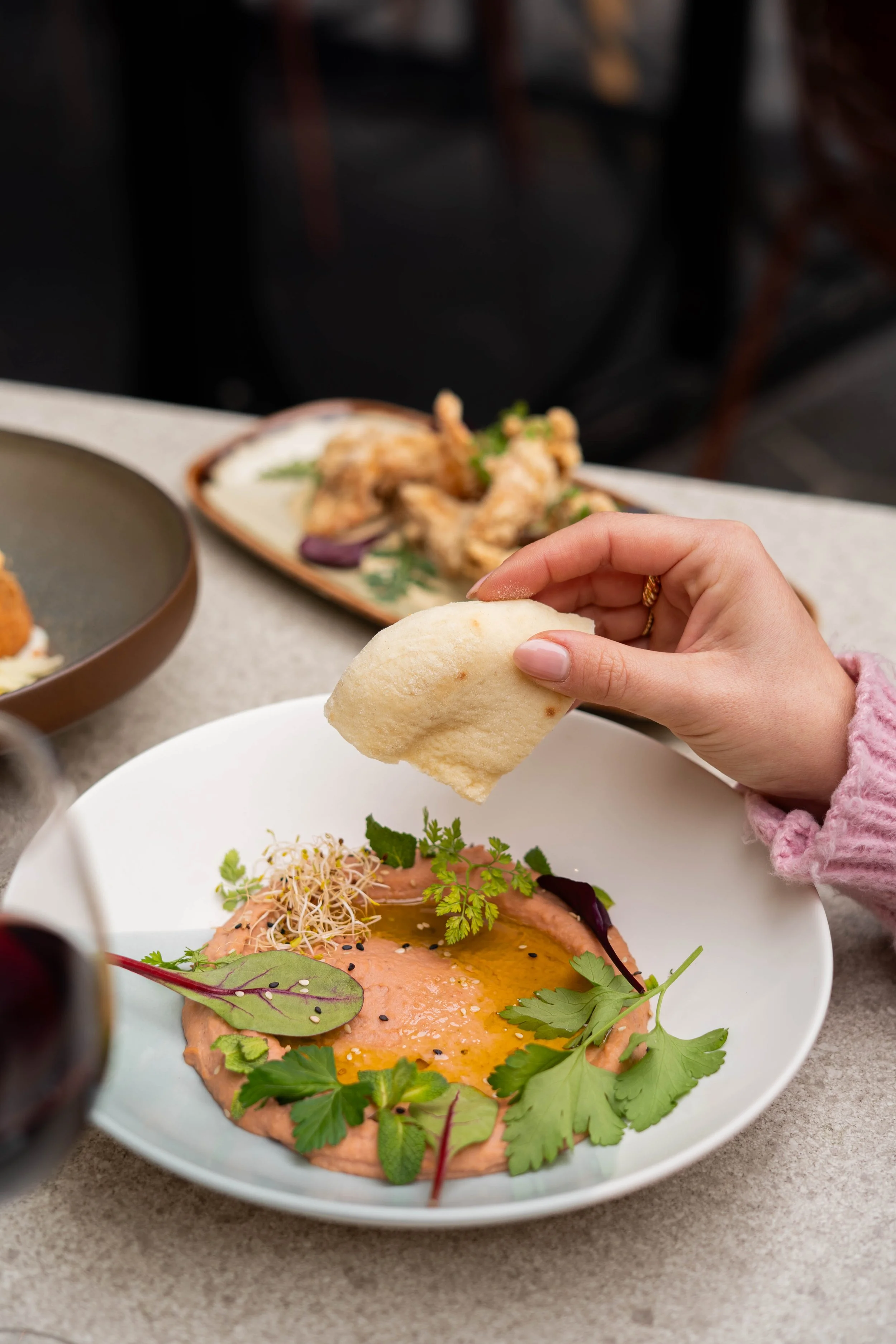 Une main tenant un morceau de pain pour tremper dans une assiette de houmous garni de feuilles de coriandre, de microgreens et d'huile.