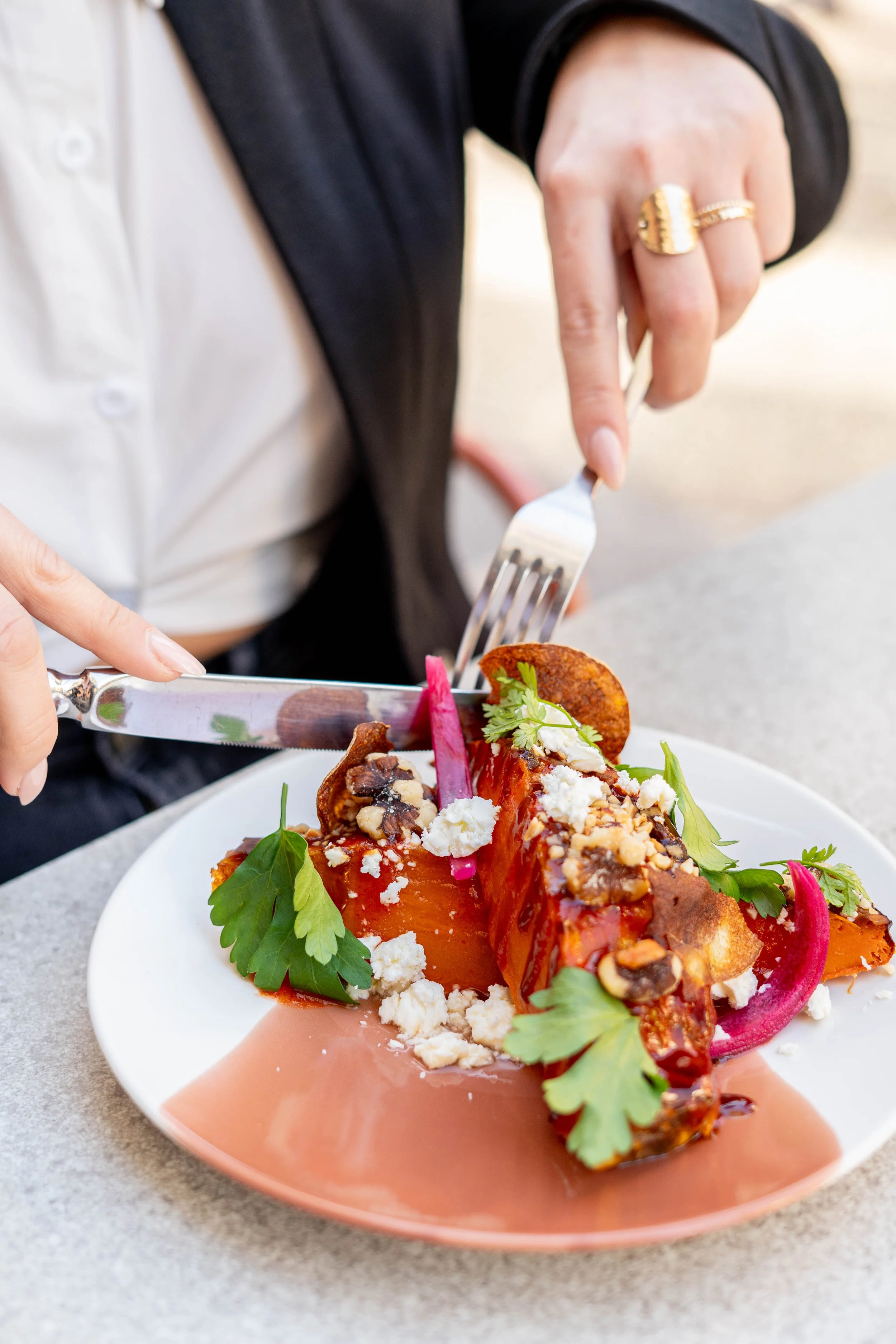 Person en train de couper une assiette de légumes grillés garnie de fromage, de noix et de feuilles de coriandre, avec une fourchette et un couteau.