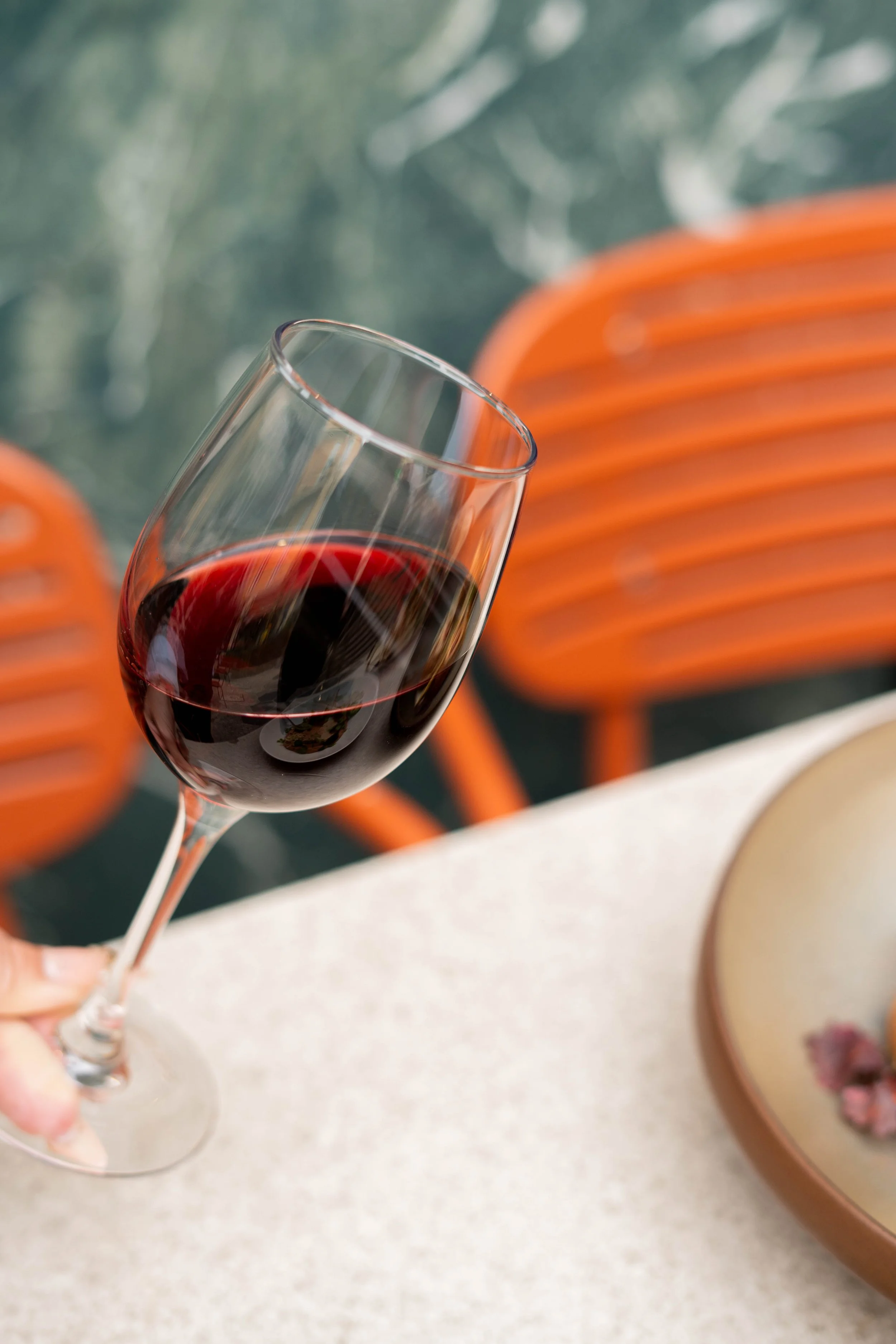 Verre de vin rouge tenu par une main, avec des chaises orange en arrière-plan.
