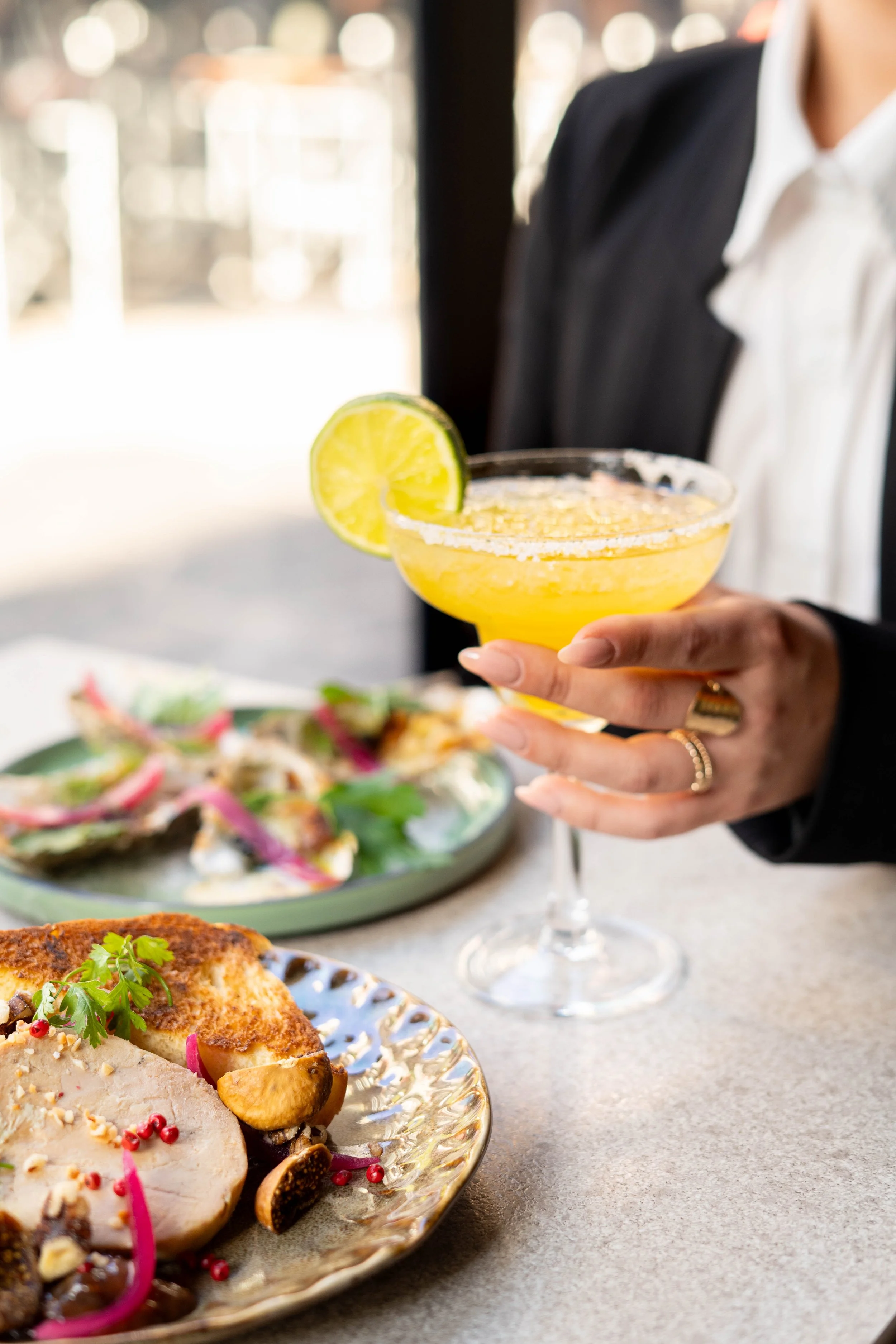 Une personne tient un verre à cocktail jaune avec une tranche de citron, devant un plat de nourriture avec viande et légumes, dans un restaurant
