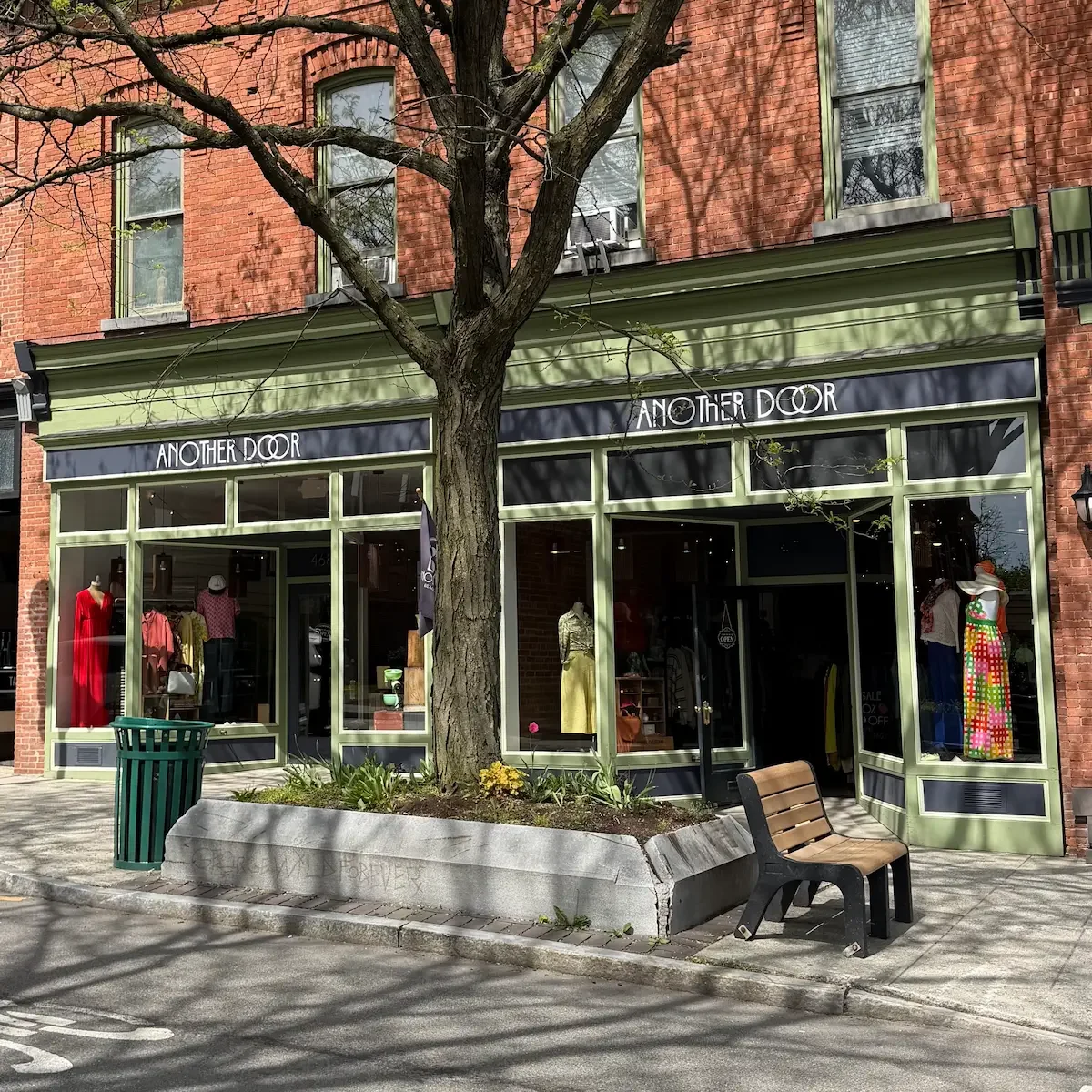Exterior of a boutique store named 'Another Door' with large glass windows, green trim, and a door with a neon 'Open' sign. A flag bearing the store's logo is outside.