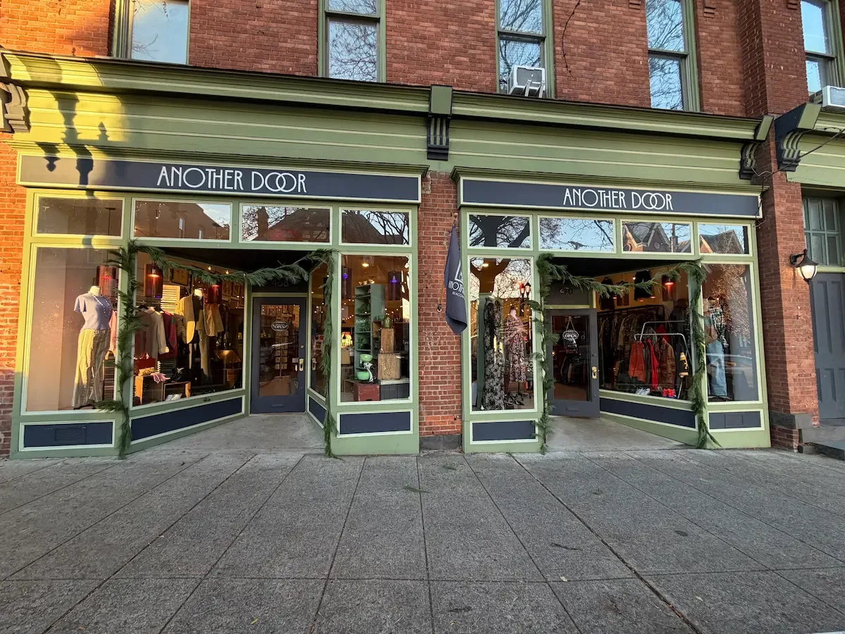 Exterior of a boutique store named 'Another Door' with large glass windows, green trim, and a door with a neon 'Open' sign. A flag bearing the store's logo is outside.