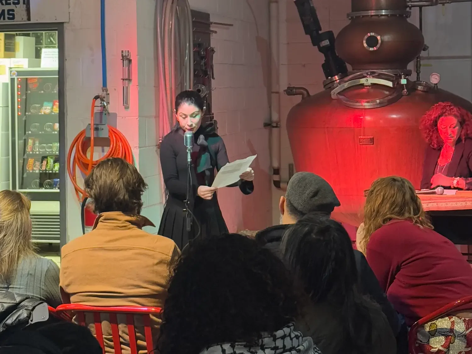 a woman reads from a script in front of an audience in a distillery