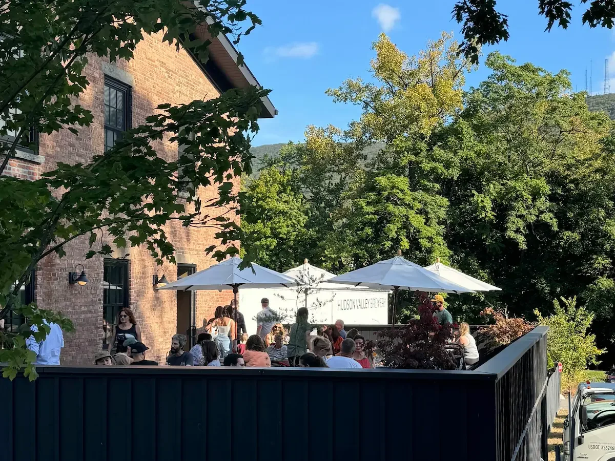 Group of people eating and drinking outside Hudson Valley Brewery in Beacon on a sunny summer day. They are surrounded by trees and shielded from the sun by white garden umbrellas