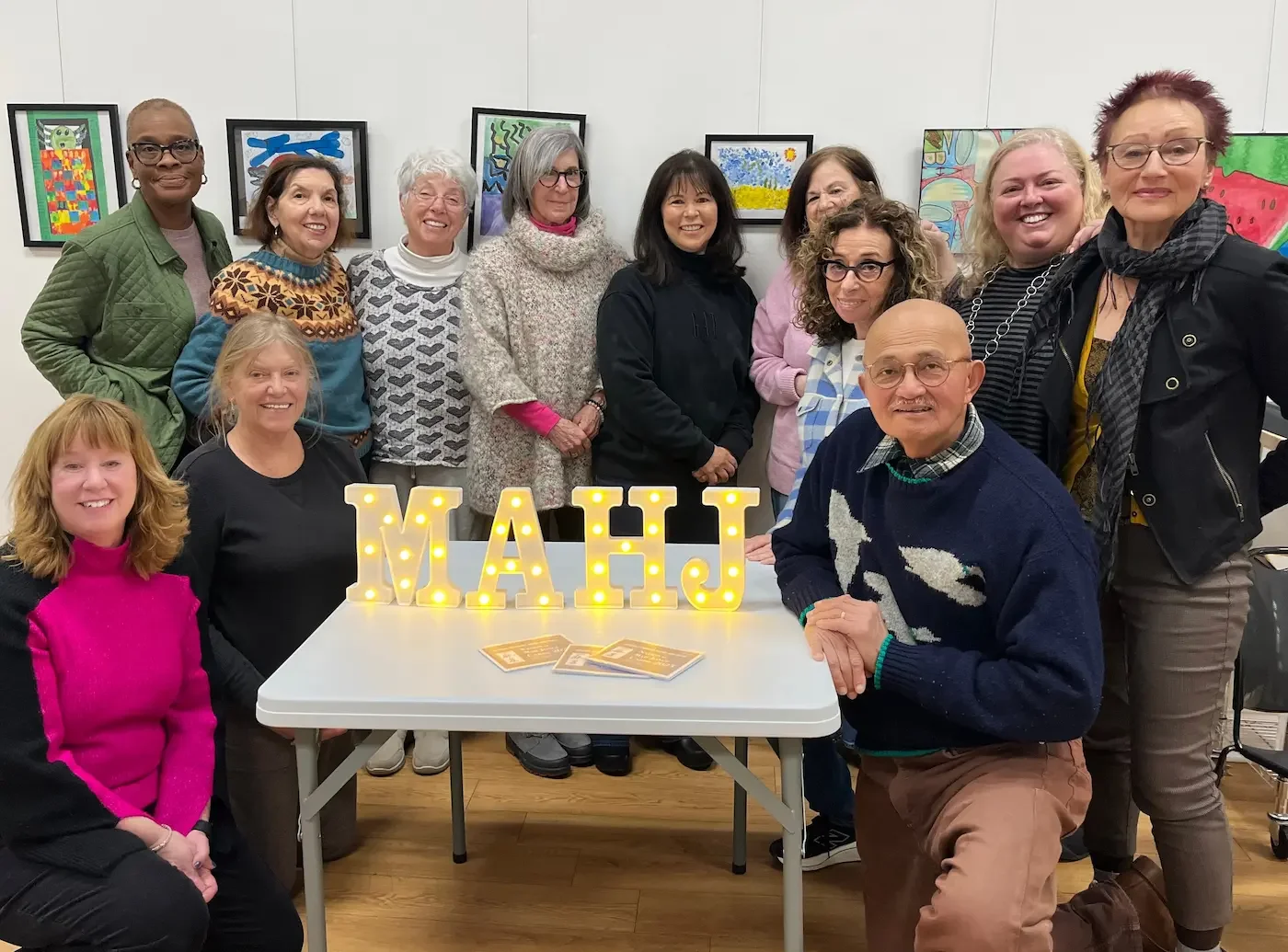 a mixed group of adults sit and stand posing for a photo with the letters MAHJ on the table