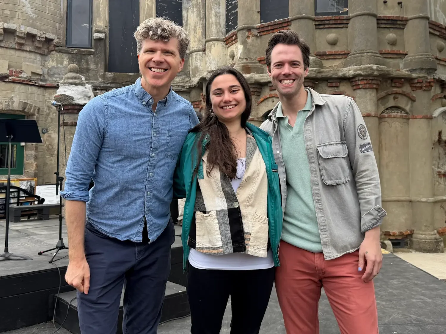 Three performers stand in front of an outdoor stage and a historic mansion, smiling for the camera.