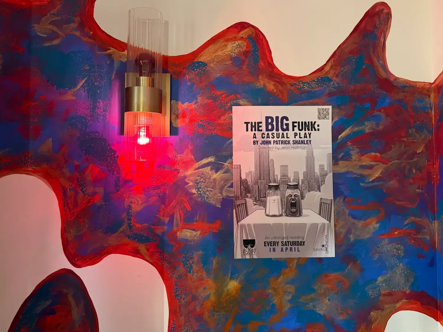 a flyer for a play called The Big Funk is posted on a wall that's splashed with reds, blues, and yellows. A wall scounce with a red bulb sits next to the flyer.