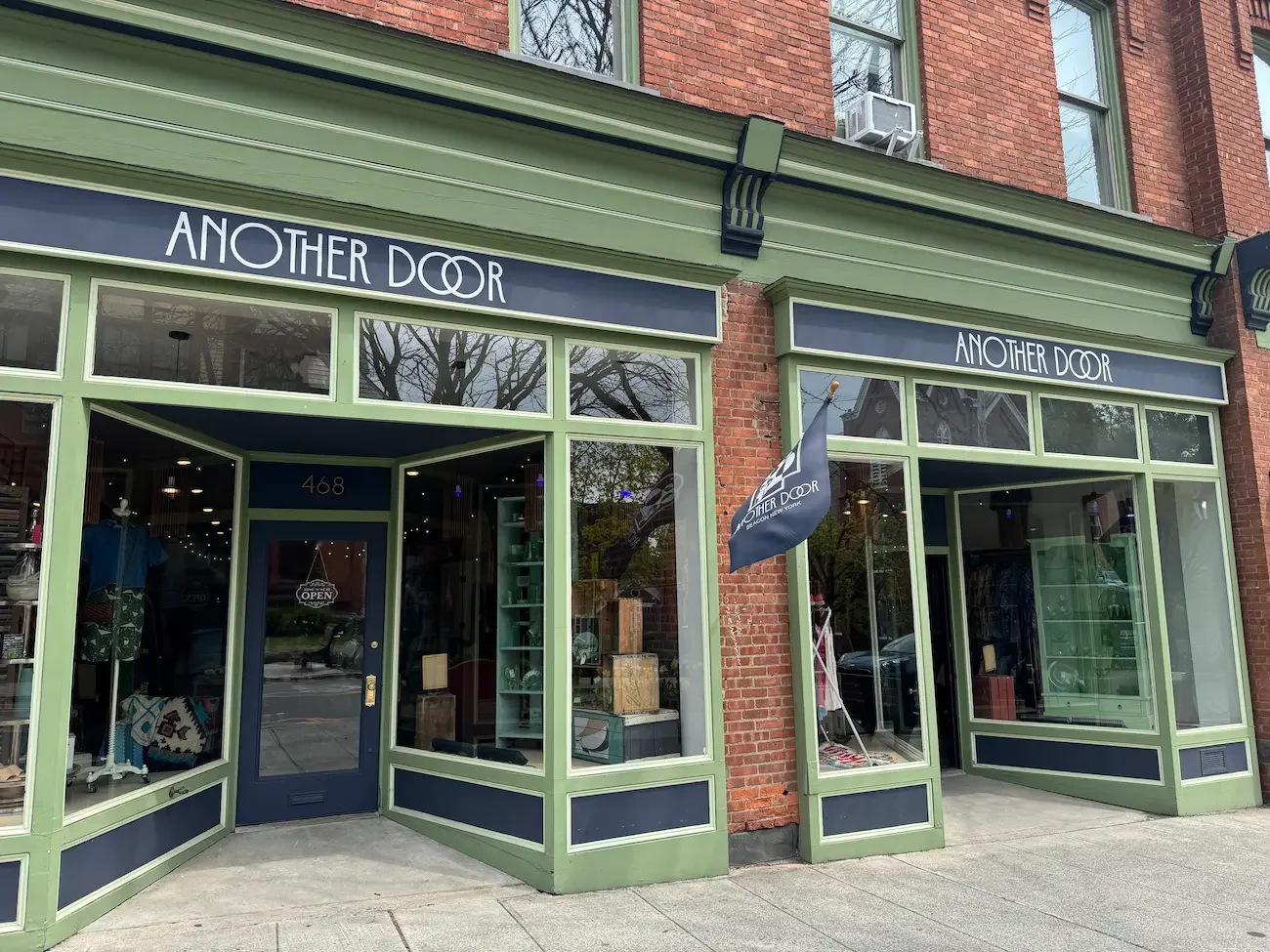 Exterior of a boutique store named 'Another Door' with large glass windows, green trim, and a door with a neon 'Open' sign. A flag bearing the store's logo is outside.