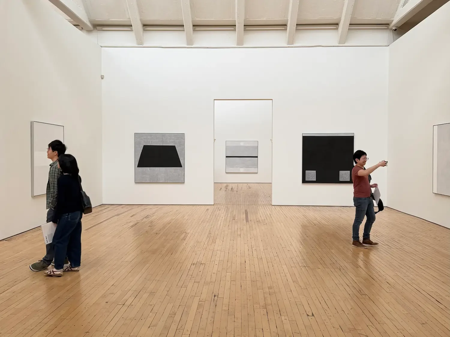 Three people in Dia Beacon art gallery viewing an exhibition by Agnes Martin. The paintings are abstract in shades of white, gray, and black. One person is taking a photo of the work.