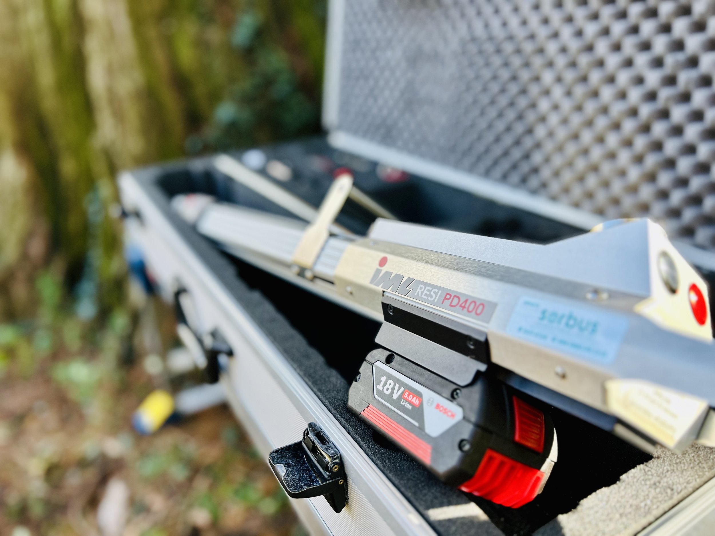 Close-up of a the Specialist IML Resi PD400 microdrill used to assess internal decay and wood strength in trees, with a blurred background of green and brown foliage.