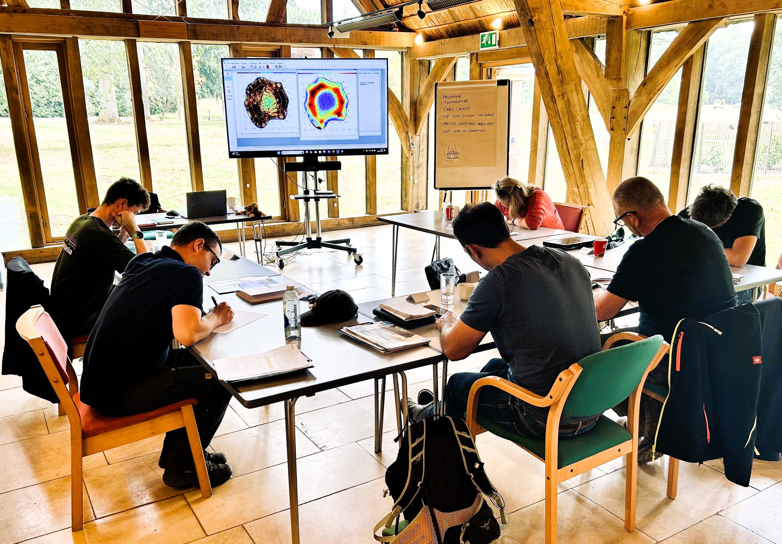 A group of people sitting in a classroom style setting taking notes in a workshop inside a wooden room with large windos. A large monitor displays colourful scientific graphs showing , and a whiteboard with handwritten notes is nearby.