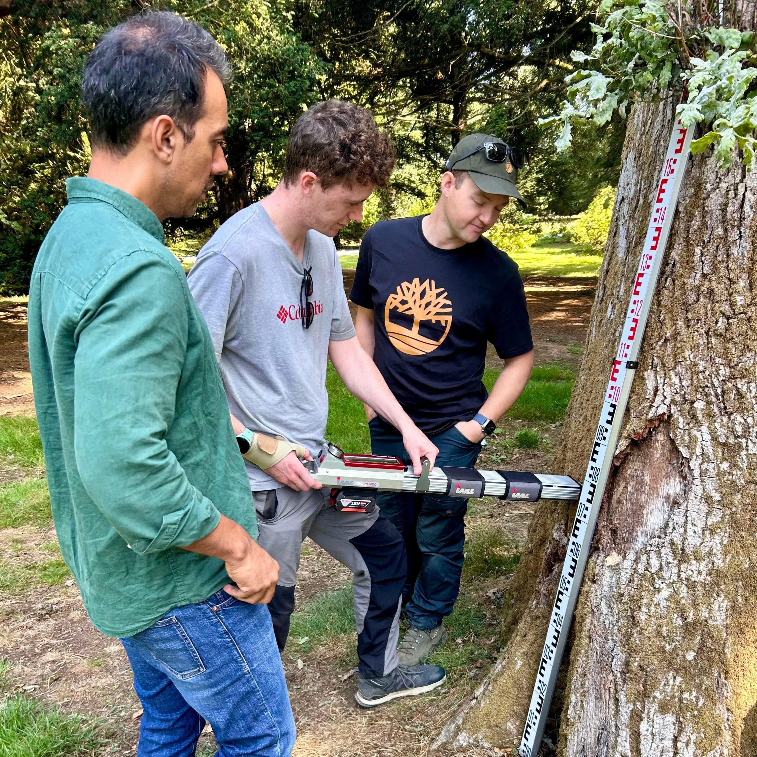 Men standing outdoors near a tree, using the IML Resi-PD400 Microdrill on the tree trunk. The background shows trees and greenery.