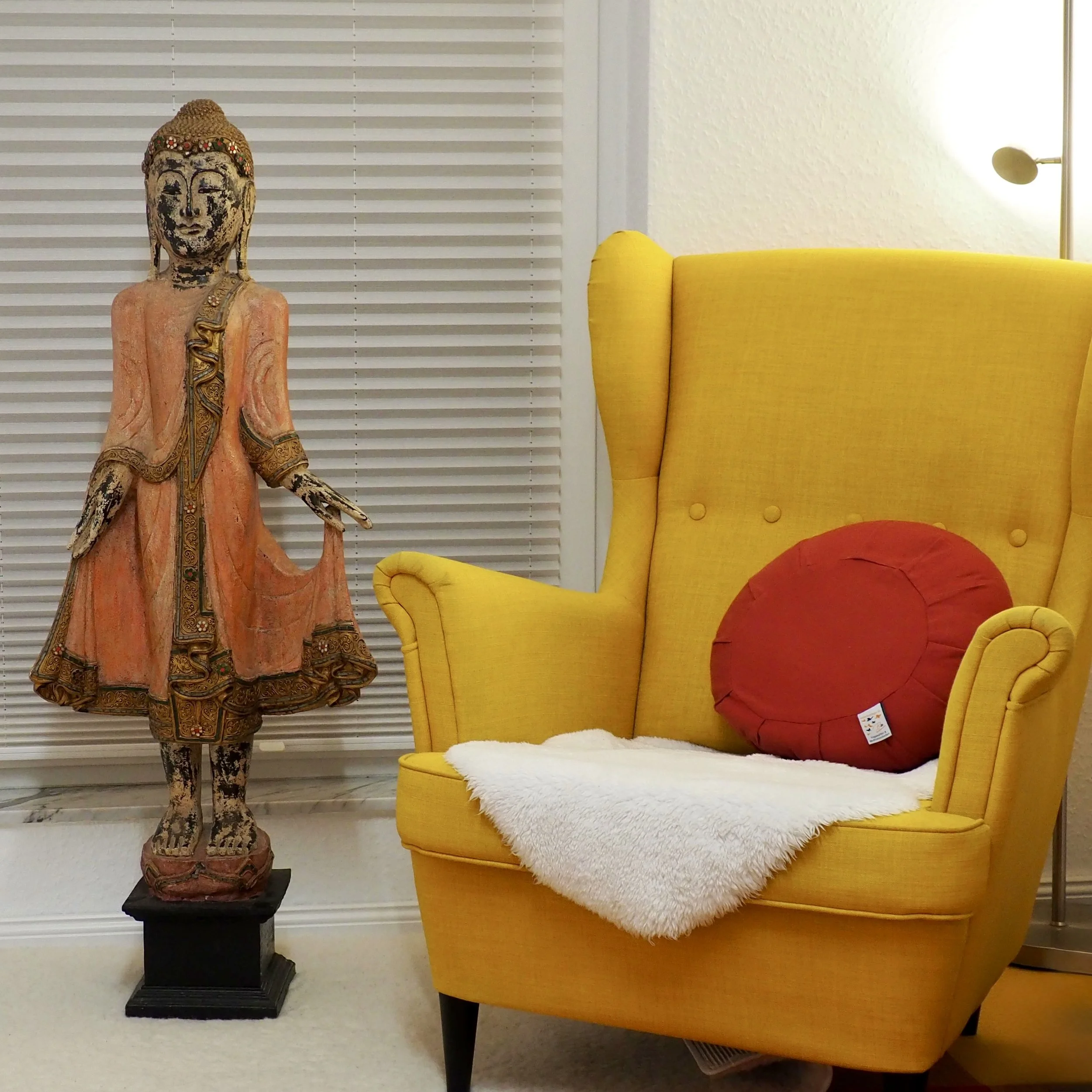 Eine geschnitzte Buddha-Figur neben einem gelben Sessel mit rotem Kissen und einer weißen Decke. Im Hintergrund sind weiße Rollos und eine weiße Wand.