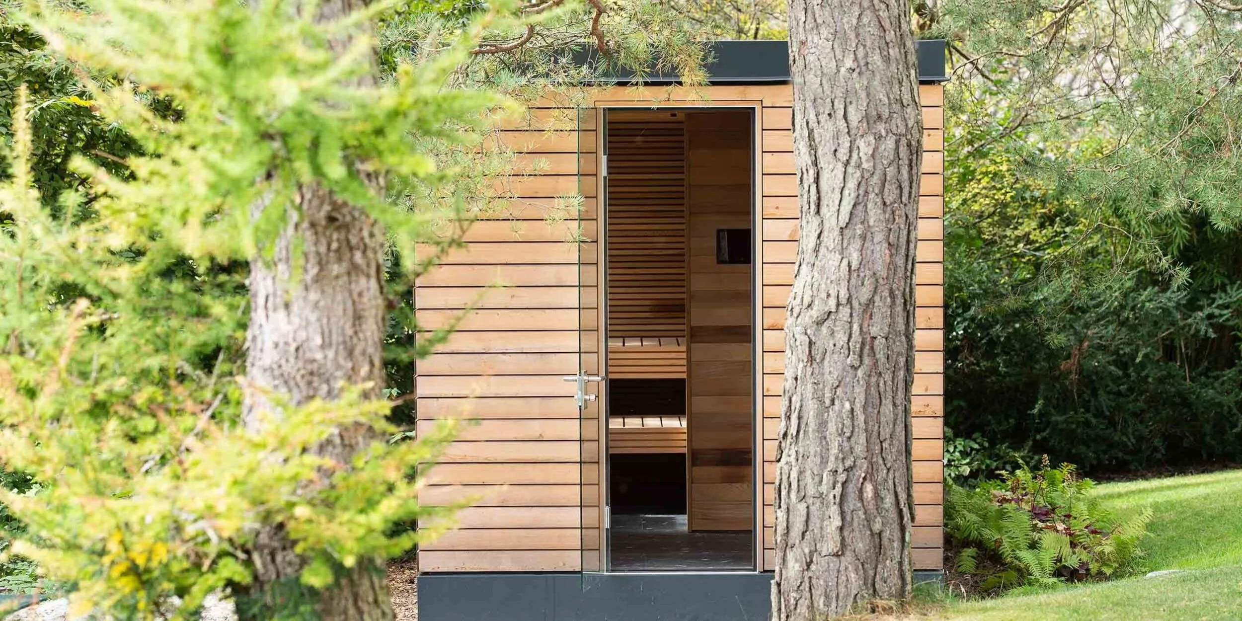 Small wooden sauna or garden house with an open door, surrounded by trees and green foliage in a garden setting.