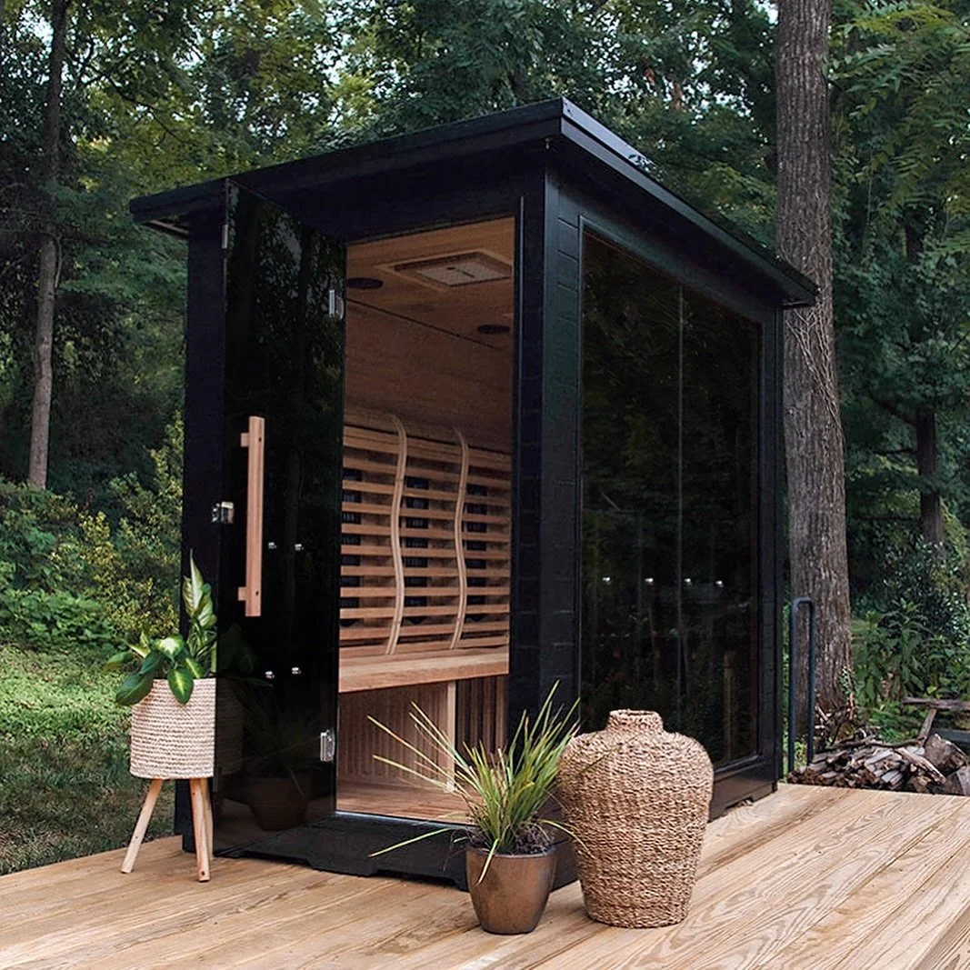 A black outdoor sauna with glass doors and wooden interior, placed on a wooden deck surrounded by trees, with potted plants and decorative objects outside.
