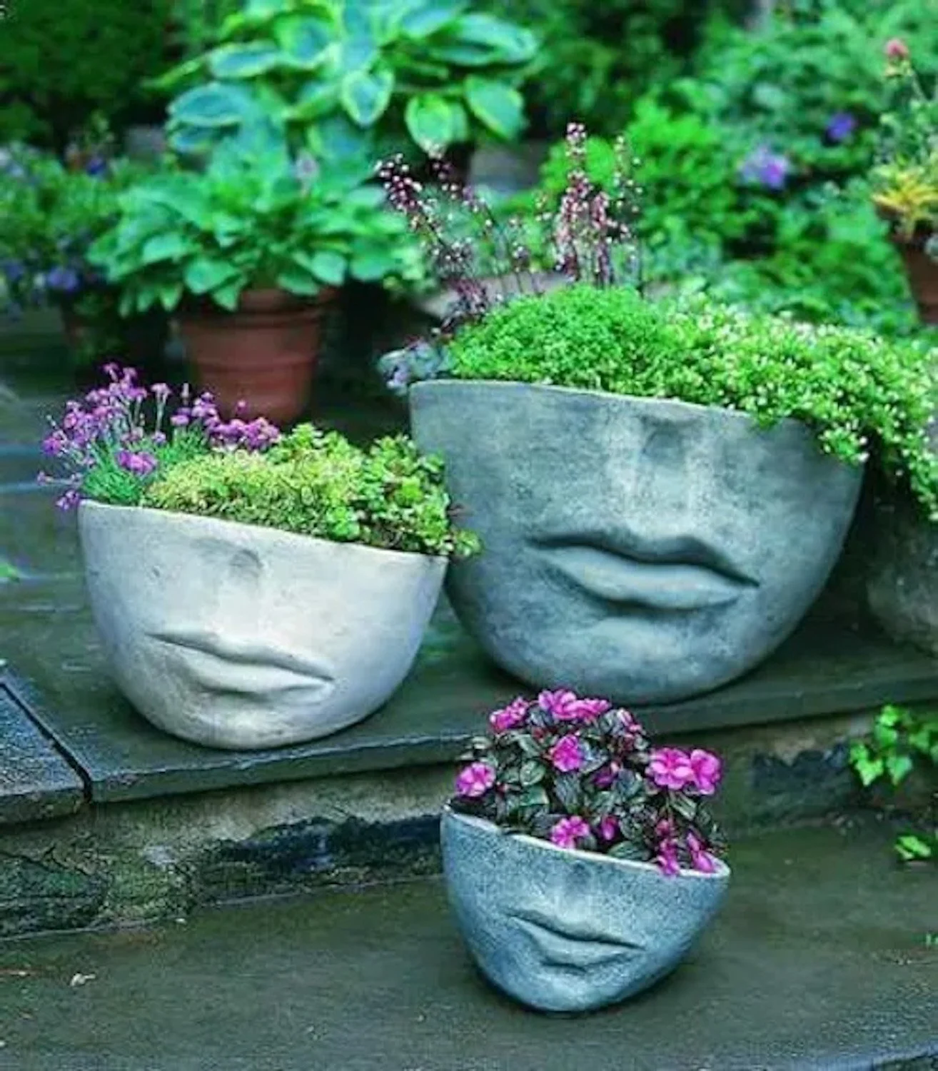 Three flower pots shaped like human faces with closed eyes and lips, filled with colorful flowers, placed on a wooden surface with potted green plants in the background.