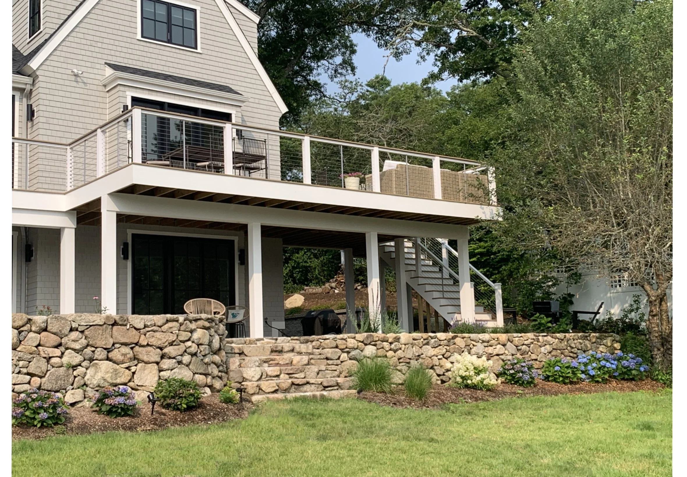 A two-story house with a large backyard featuring a stone retaining wall, a grassy lawn, and a landscaped garden with purple and white hydrangeas. The house has an upper deck with outdoor furniture and a lower covered patio with access through stairs