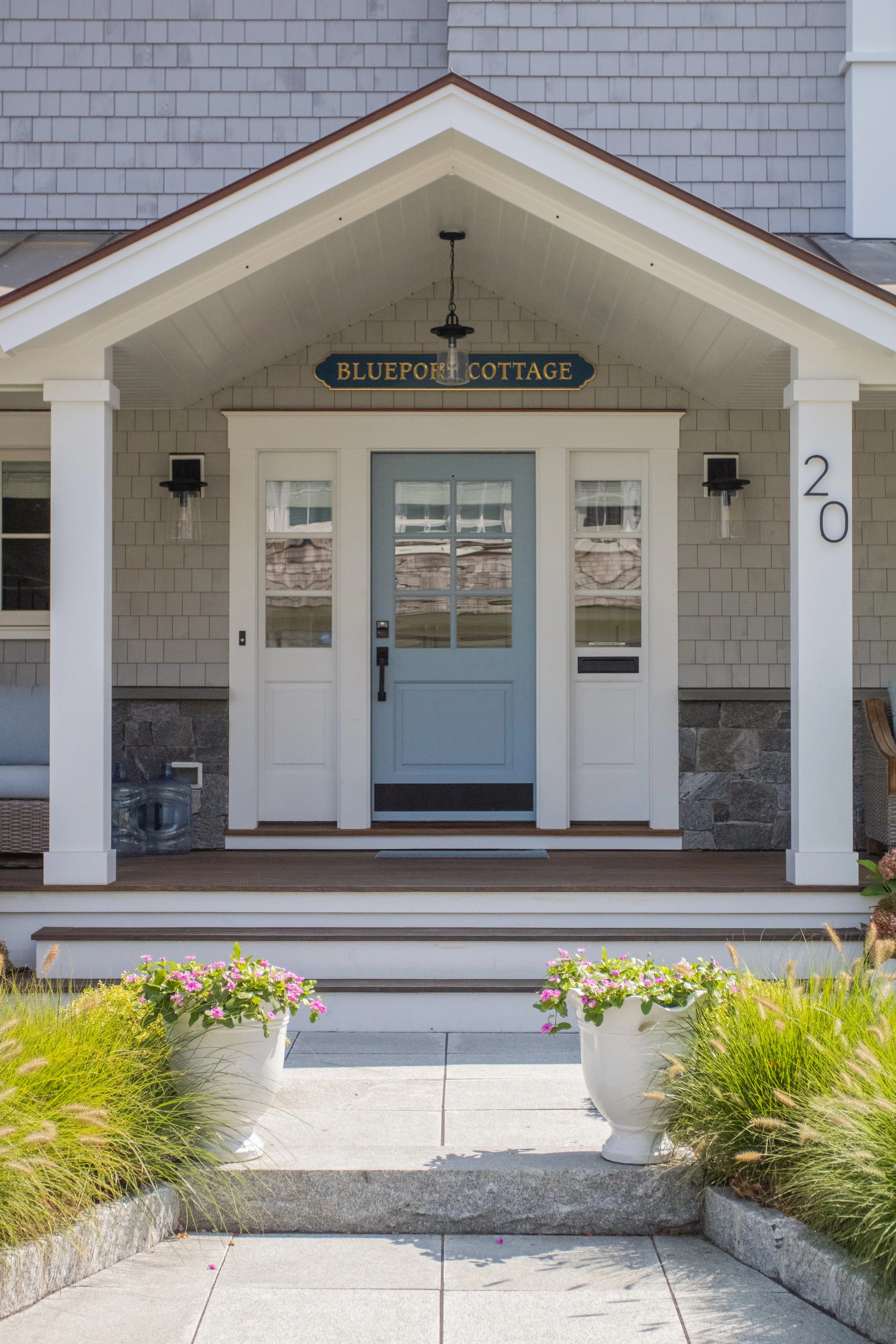 Front porch of a house with a blue door, white trim, and a sign that reads 'Blueport Cottage'. The house number 20 is on the right column, and two large white planters with pink flowers are in front of the steps leading up to the porch.