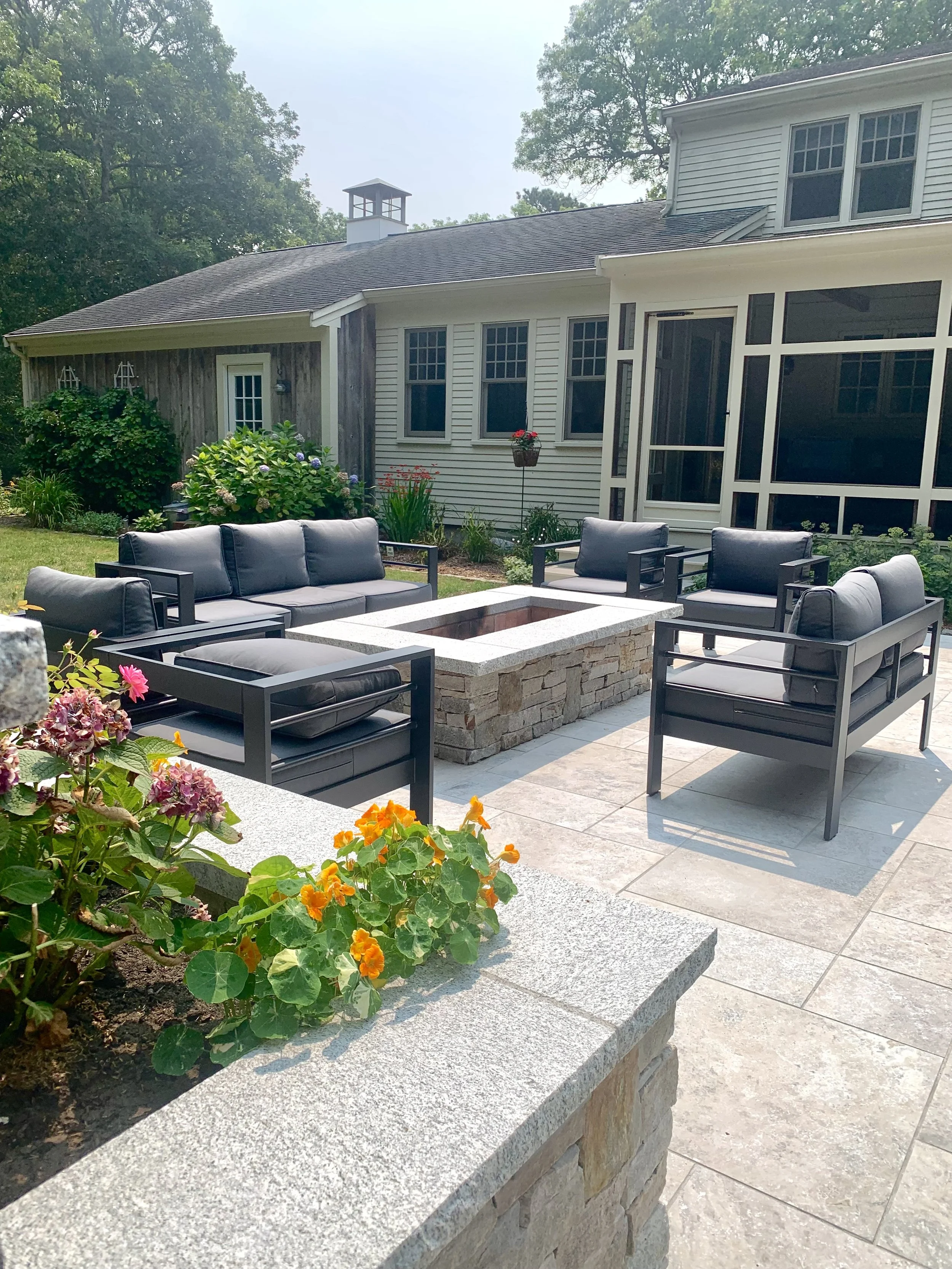 Outdoor patio with black cushioned seating arranged around a stone fire pit, surrounded by flower beds and a house with white siding and large windows.