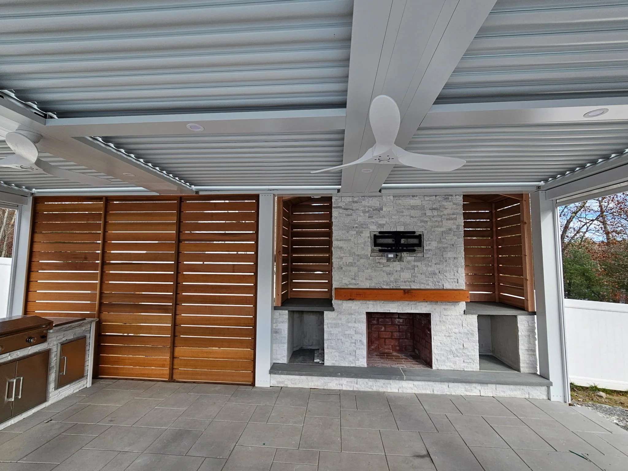 Covered outdoor patio area with a stone fireplace and ceiling fans, enclosed by wooden slat privacy screens.
