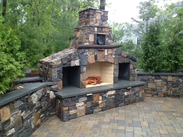 Outdoor stone fireplace with built-in firewood storage and surrounded by trees and stone wall.