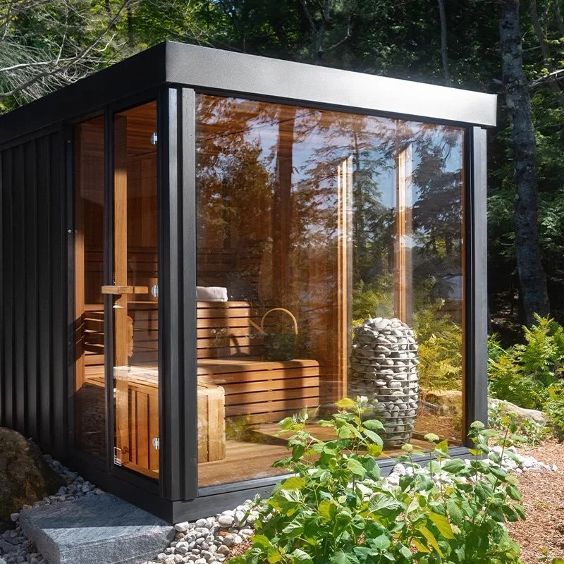 Small black metal and glass outdoor sauna with a wooden interior, surrounded by rocks and greenery.