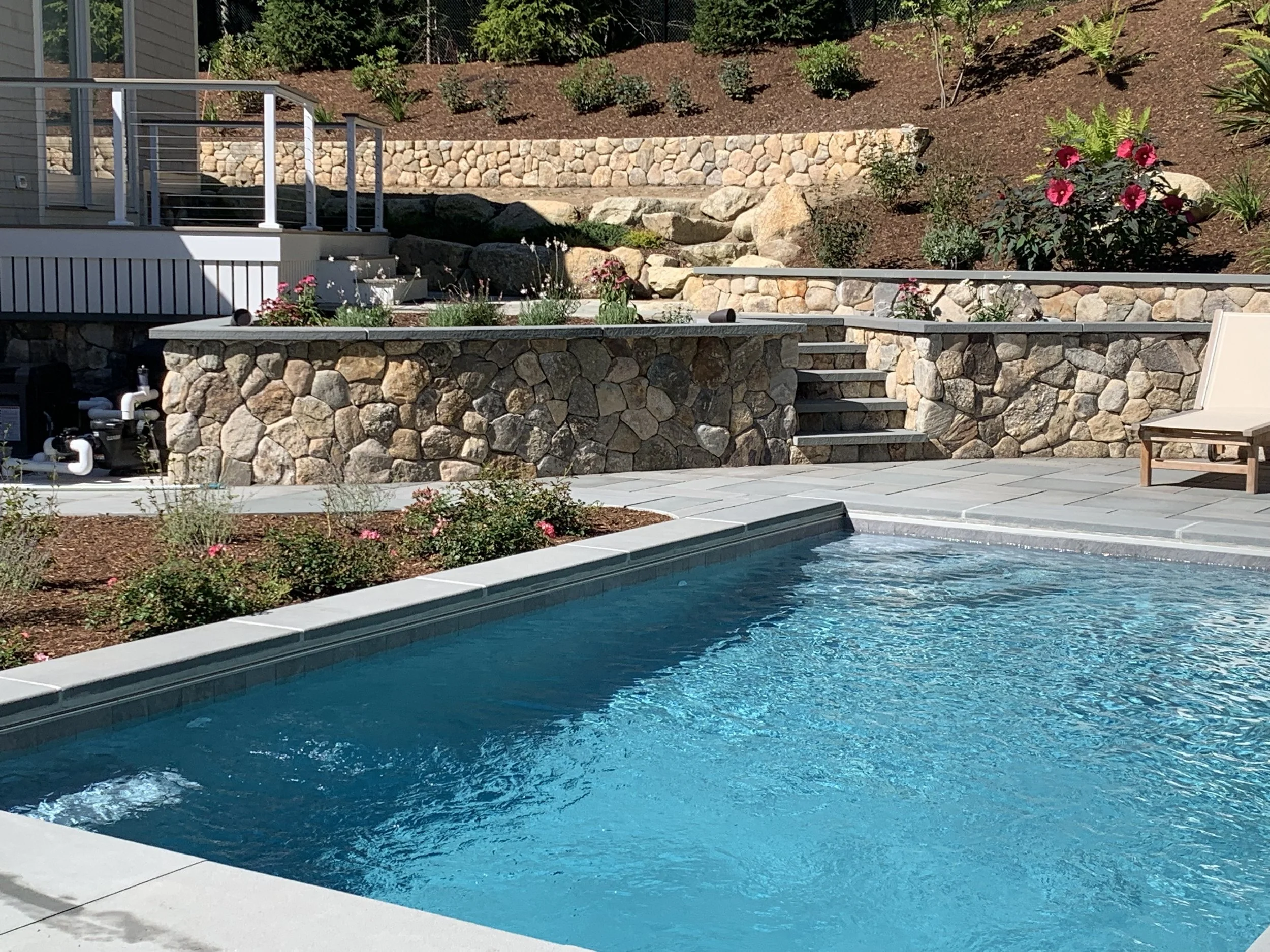 A backyard features a swimming pool with clear blue water, surrounded by gray paving stones. There are stone retaining walls with flowers and plants, and a brown chair is positioned on the patio. The background shows a hillside with small bushes and flowers.