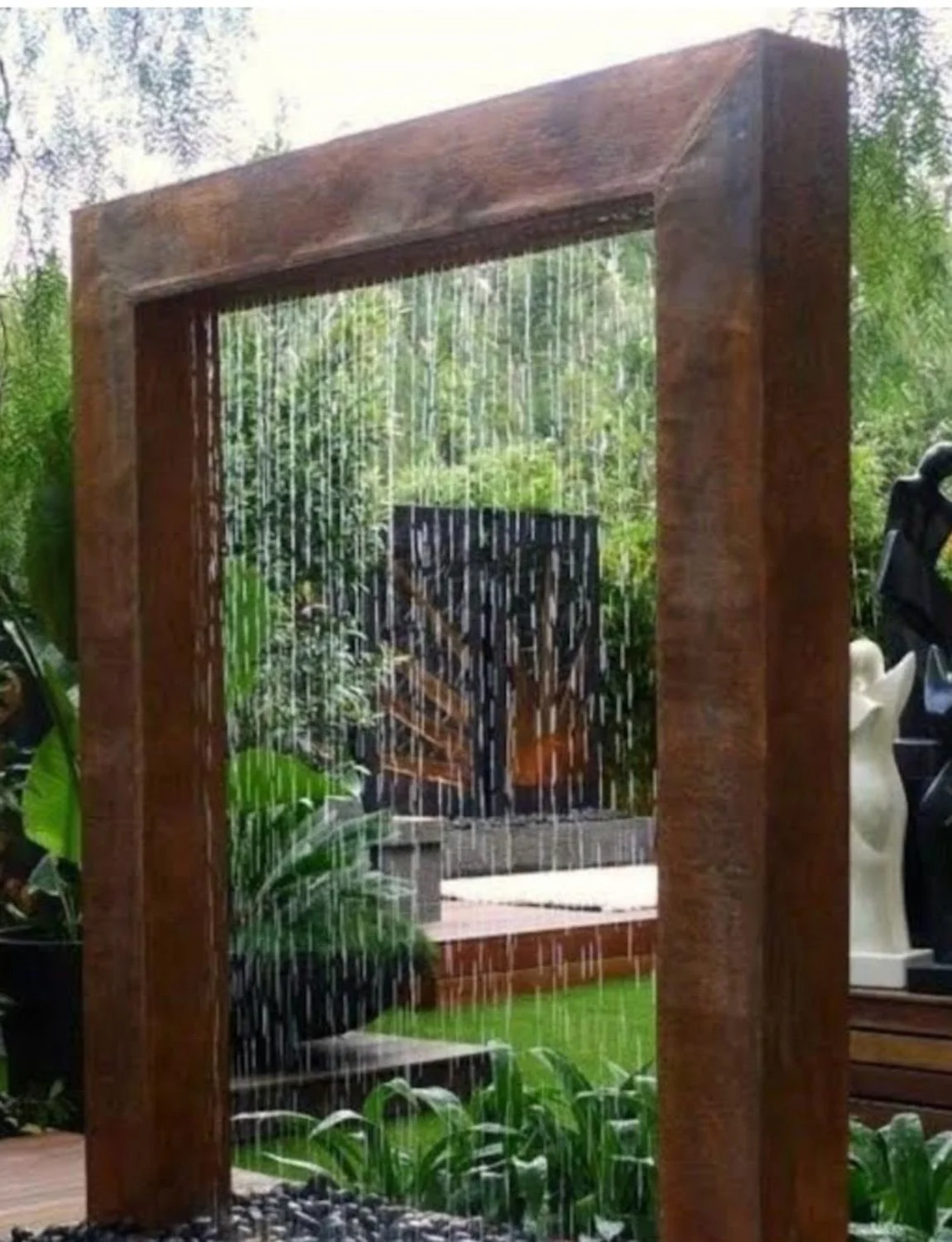 Water feature with a waterfall from a wooden frame, surrounded by lush green plants and outdoor garden decor.