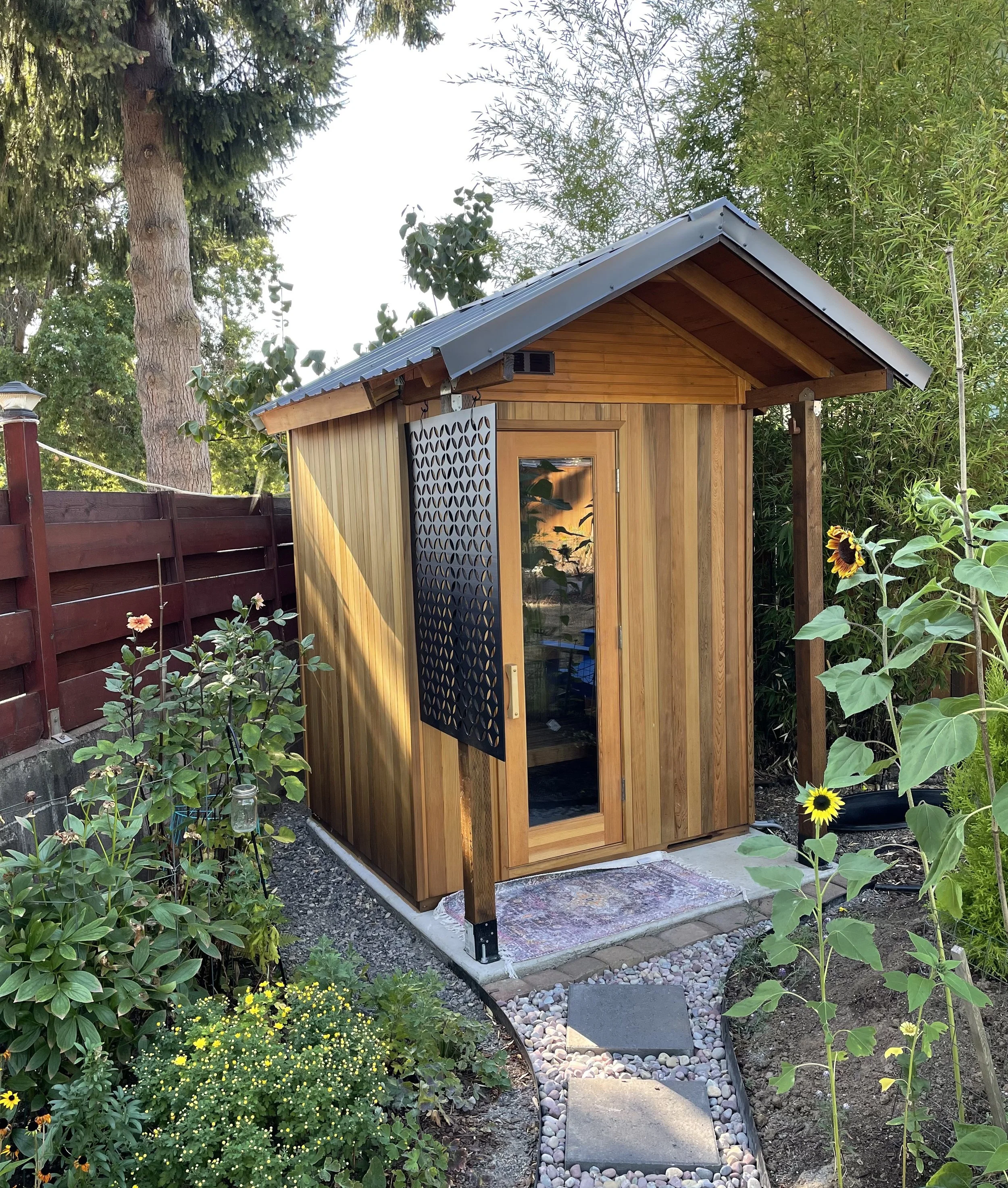 A small wooden sauna with a metal roof, a glass door, and a black decorative panel on one side, surrounded by flowers and plants in a garden.