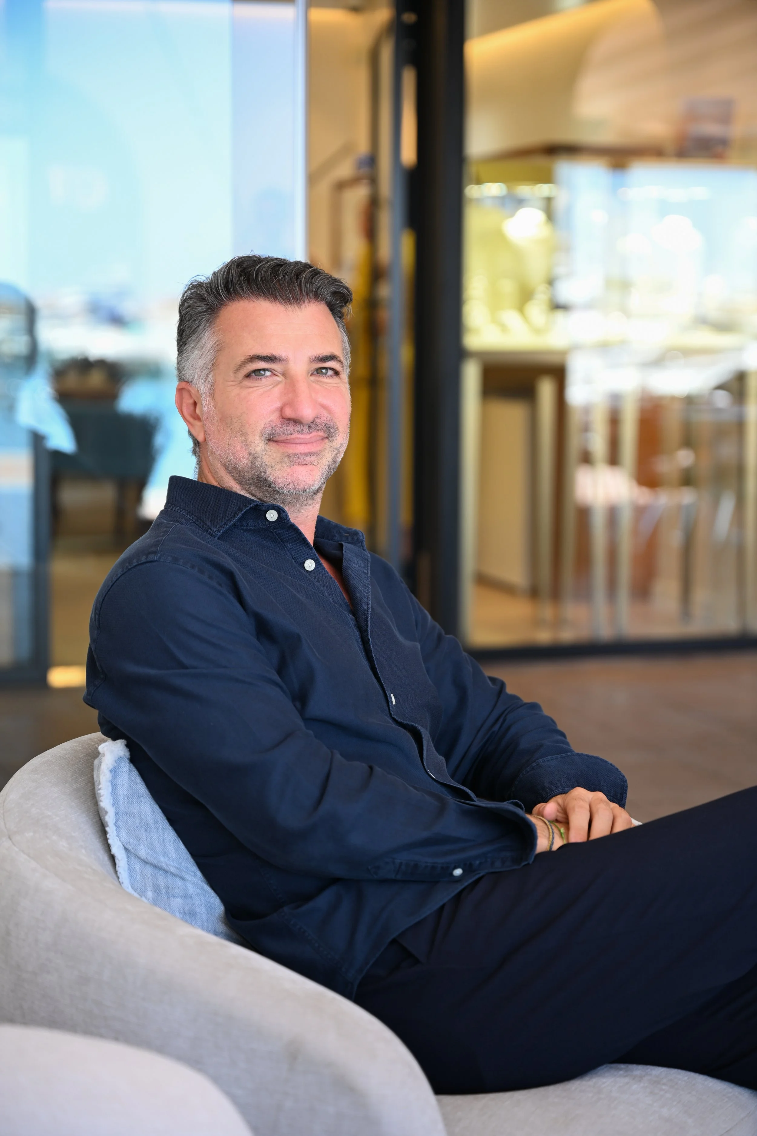 Baris Kayrak, founder of Diamotto and expert on diamonds & jewellerys in mallorca.  wearing a dark blue shirt, sitting on a light-colored armchair in a modern indoor setting, smiling at the camera. 