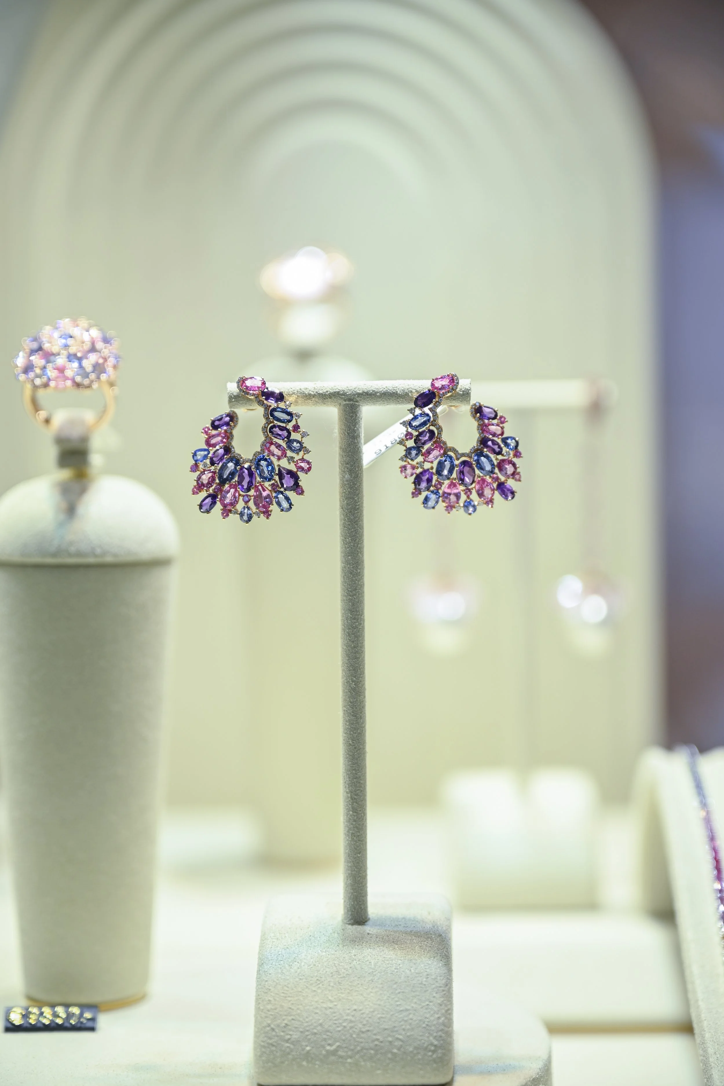 Colorful teardrop-shaped gemstone earrings displayed on a stand, with other jewelry pieces blurred in the background.