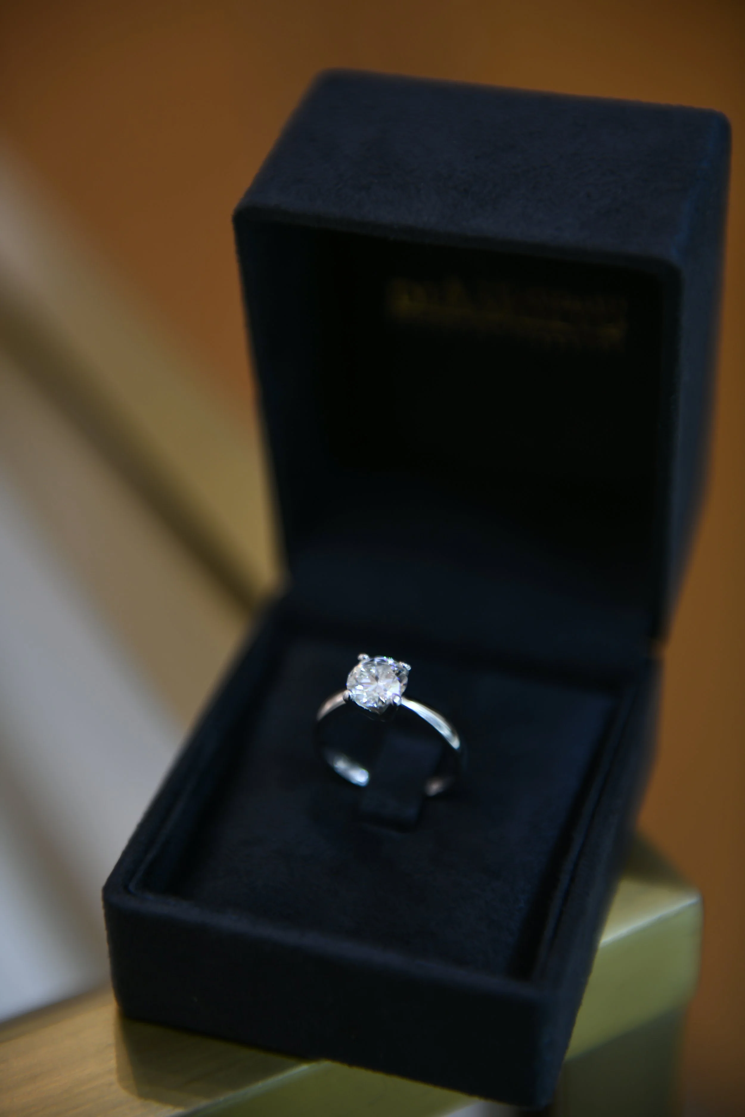 A diamond engagement ring in a black velvet ring box, sitting on a wooden surface.
