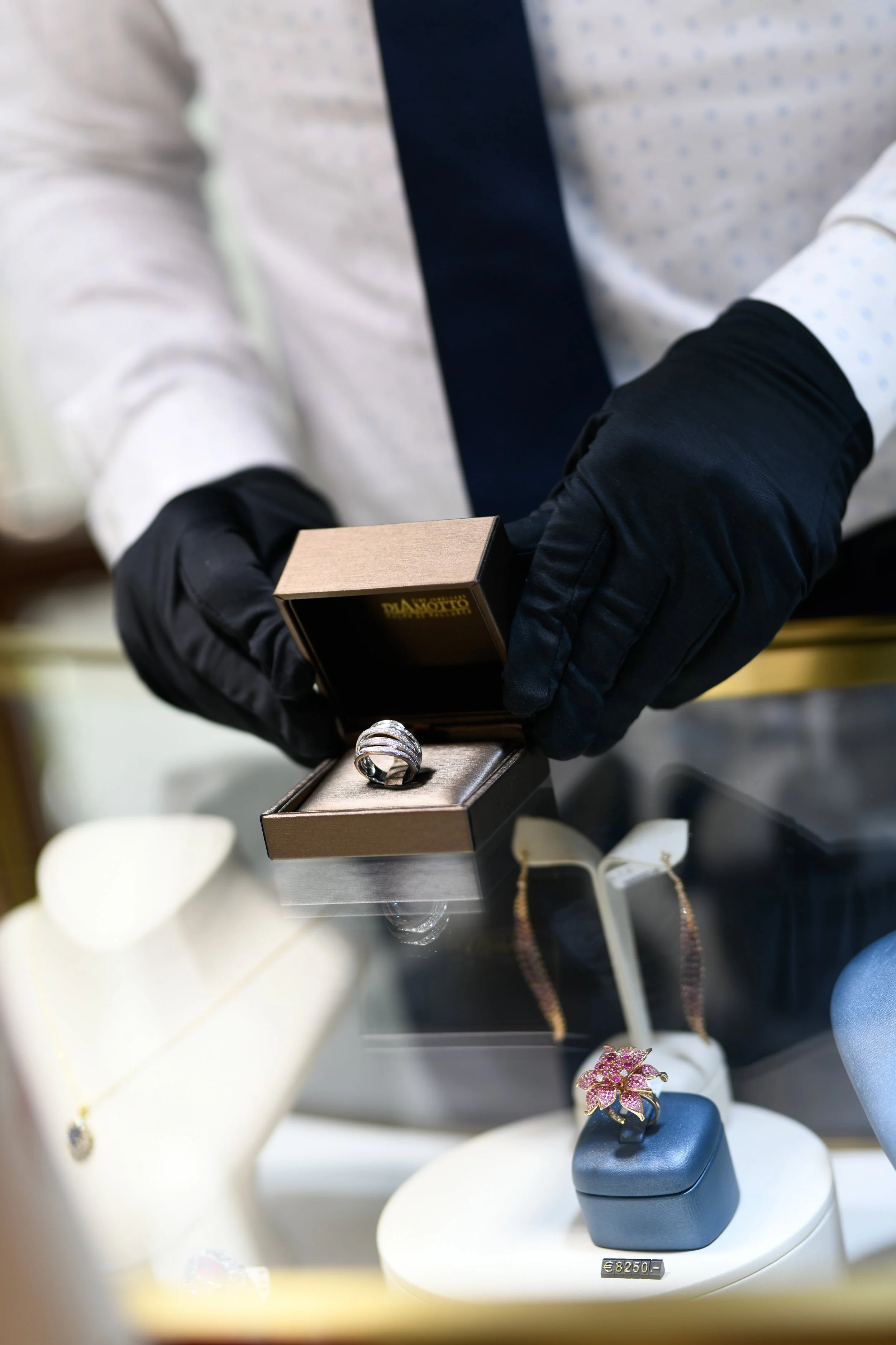 The image shows a person wearing black gloves holding an open jewelry box with a ring inside. There are various jewelry items like earrings, rings, and necklaces displayed on white stands, with a price tag of €8250 visible on one of the stands.