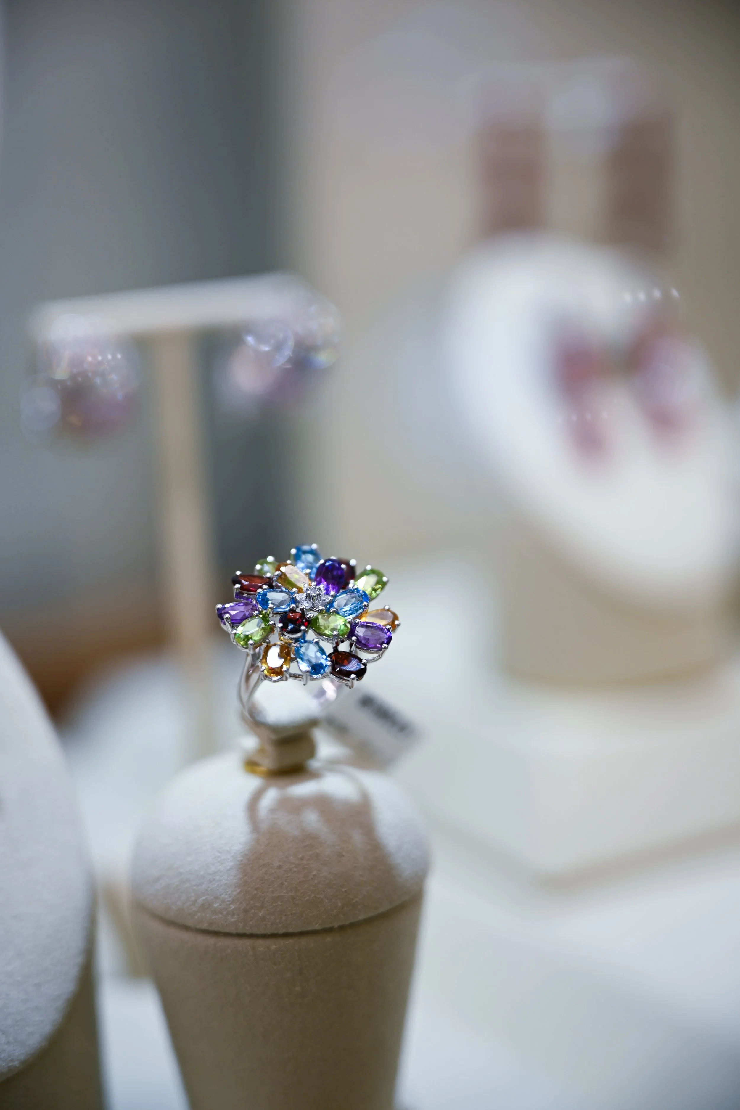 A colorful flower-shaped ring with multiple gemstones on a jewelry display stand, with a blurred background showing textiles or clothing.