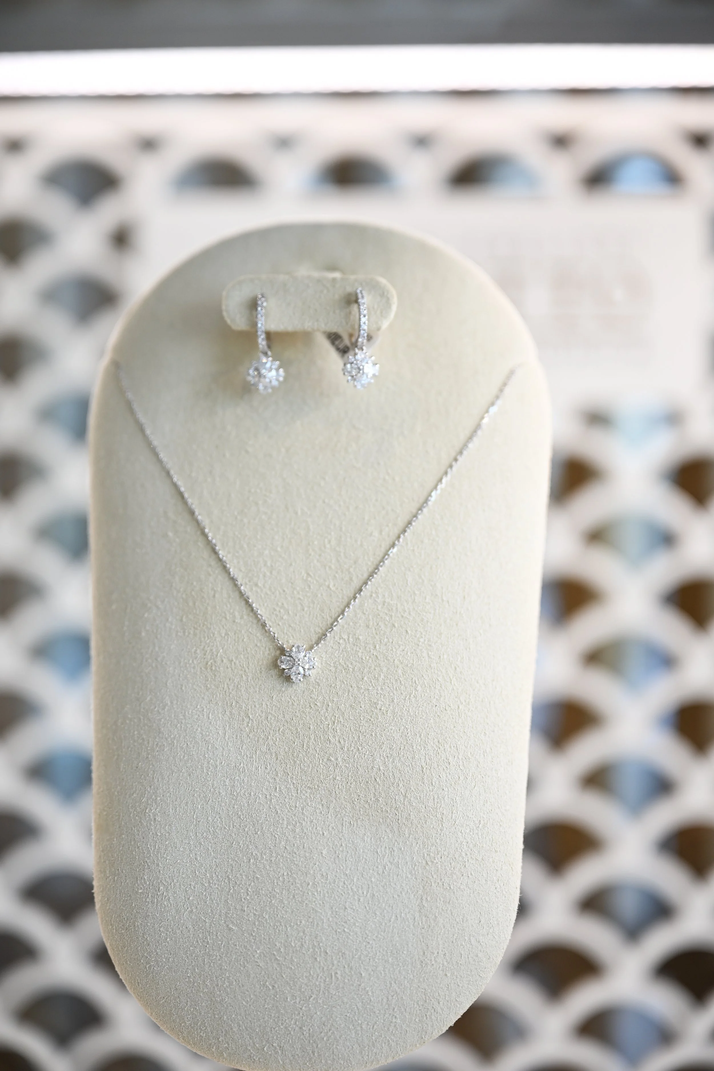 A jewelry display stand with diamond earrings in the shape of flowers and a matching necklace, in front of a blurred lattice background.