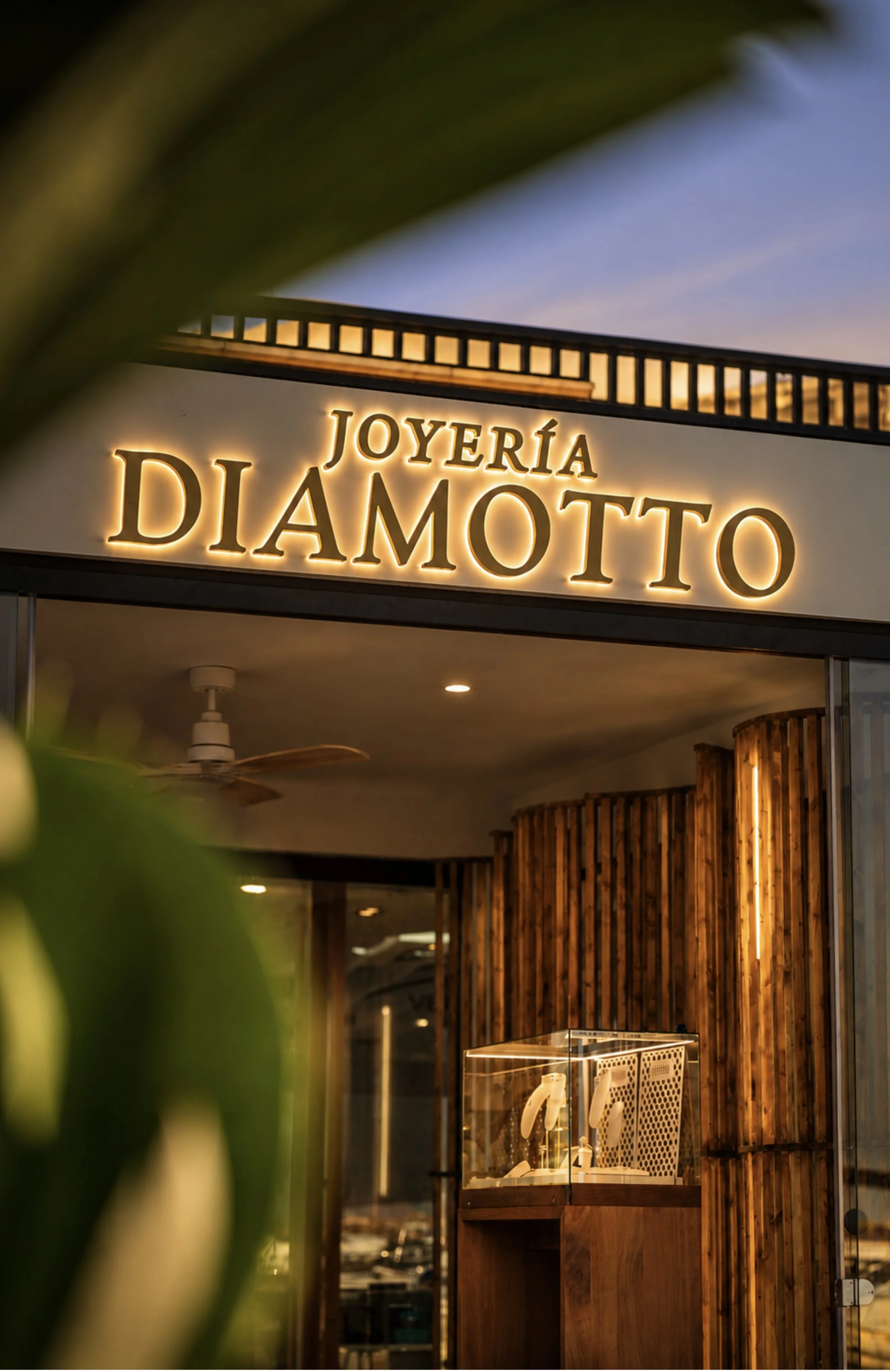 Storefront sign that reads "Joyería Damotto" illuminated at night, with interior lighting visible through large windows, wooden paneling, and decorative elements inside.